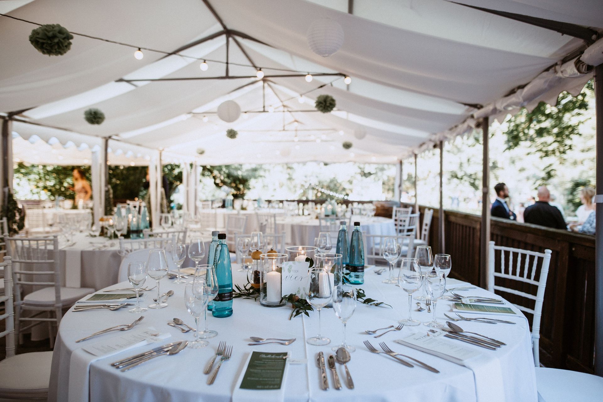 Hochzeitsempfangstische unter einem weißen Zelt, dekoriert mit Kerzen und Grünpflanzen.