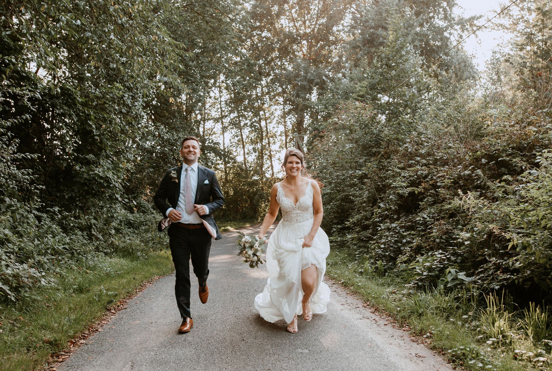 Frischvermählte laufen freudig einen von Bäumen gesäumten Weg entlang; die Braut hält einen Blumenstrauß und beide lächeln.