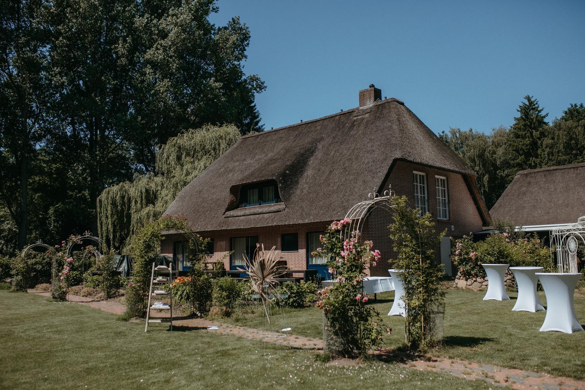 Reetgedecktes Haus mit Garten; weiße Cocktailtische auf dem Rasen, sonniger Tag.