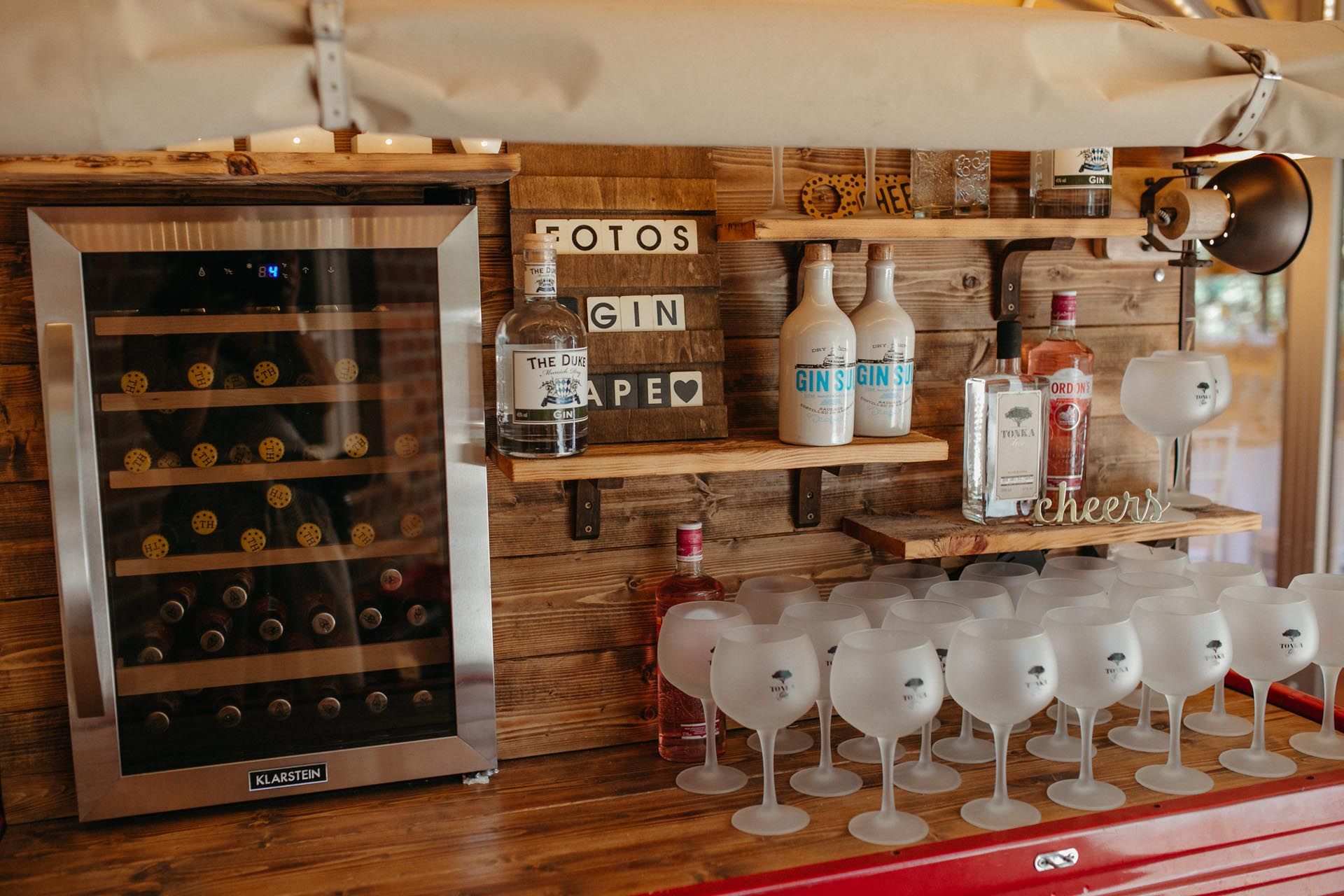 Weinkühler, Schnapsflaschen, Glaswaren und ein Schild mit der Aufschrift „Gin Grape“ auf einer Holzbar.