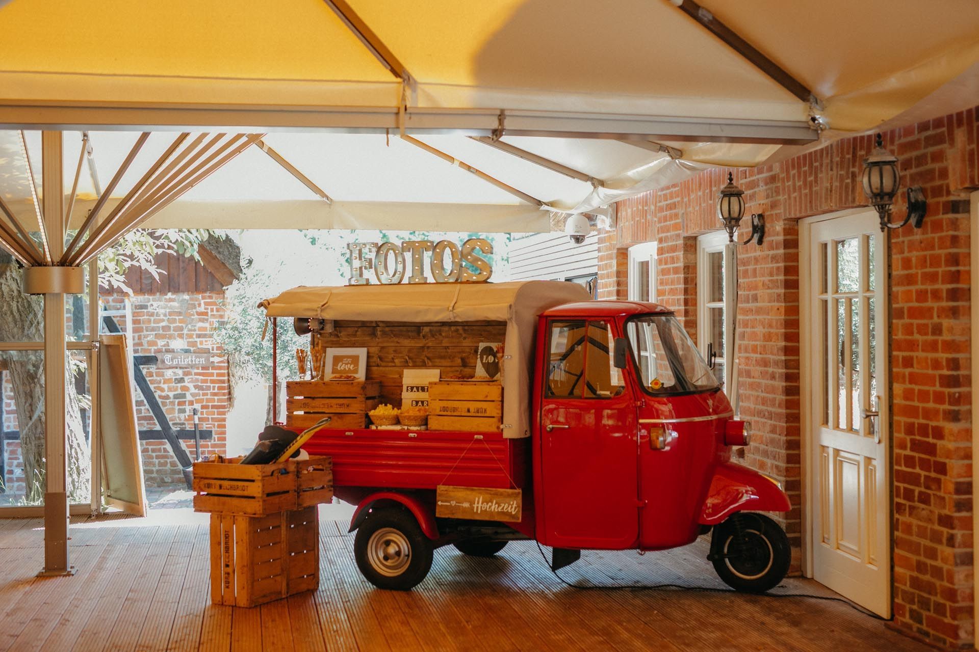 Roter Piaggio Ape-Imbisswagen, geparkt in einem Backsteingebäude mit überdachter Terrasse.
