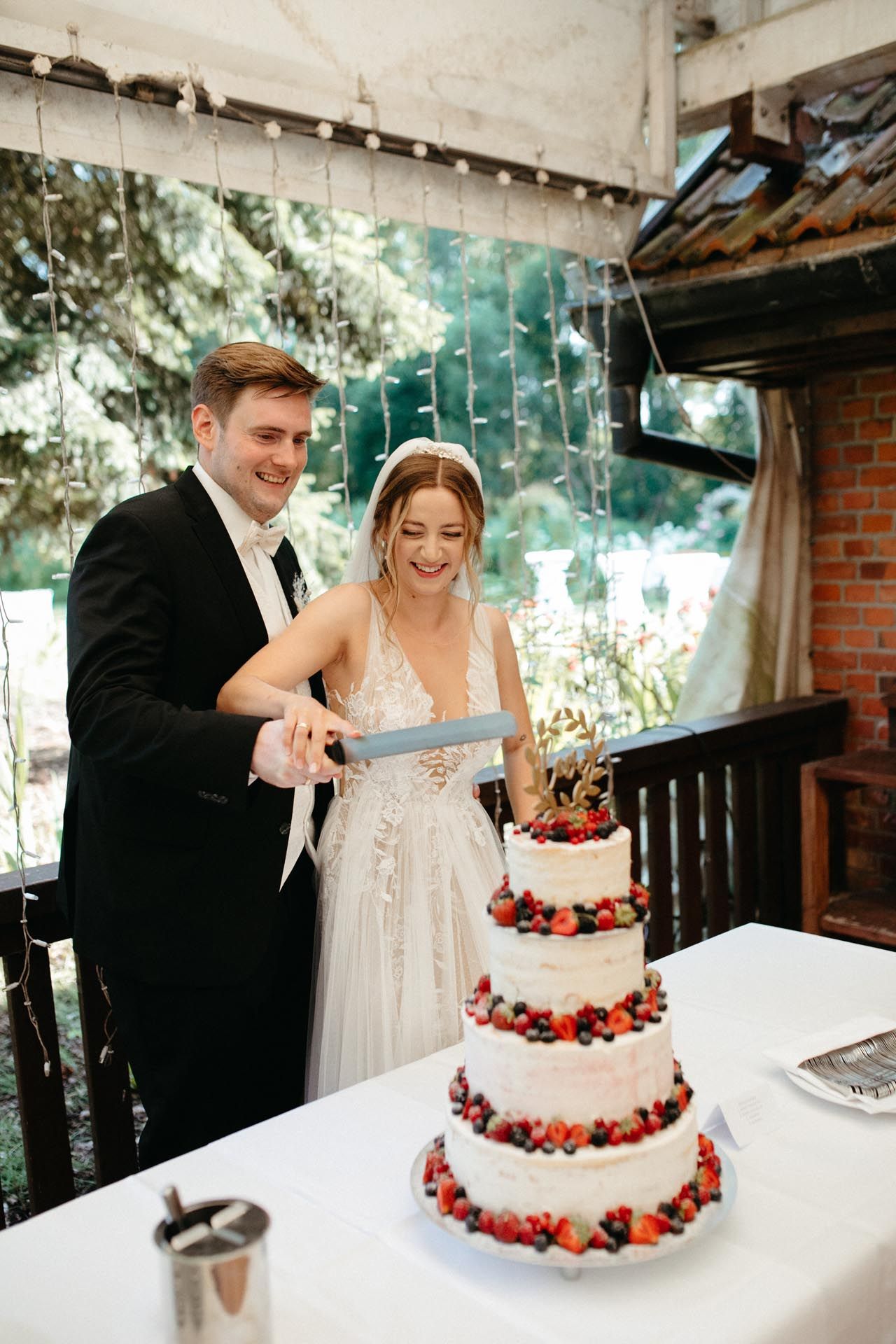 Braut und Bräutigam schneiden eine mehrstöckige Hochzeitstorte an, die mit Beeren dekoriert ist. Lächelndes Paar im Freien.