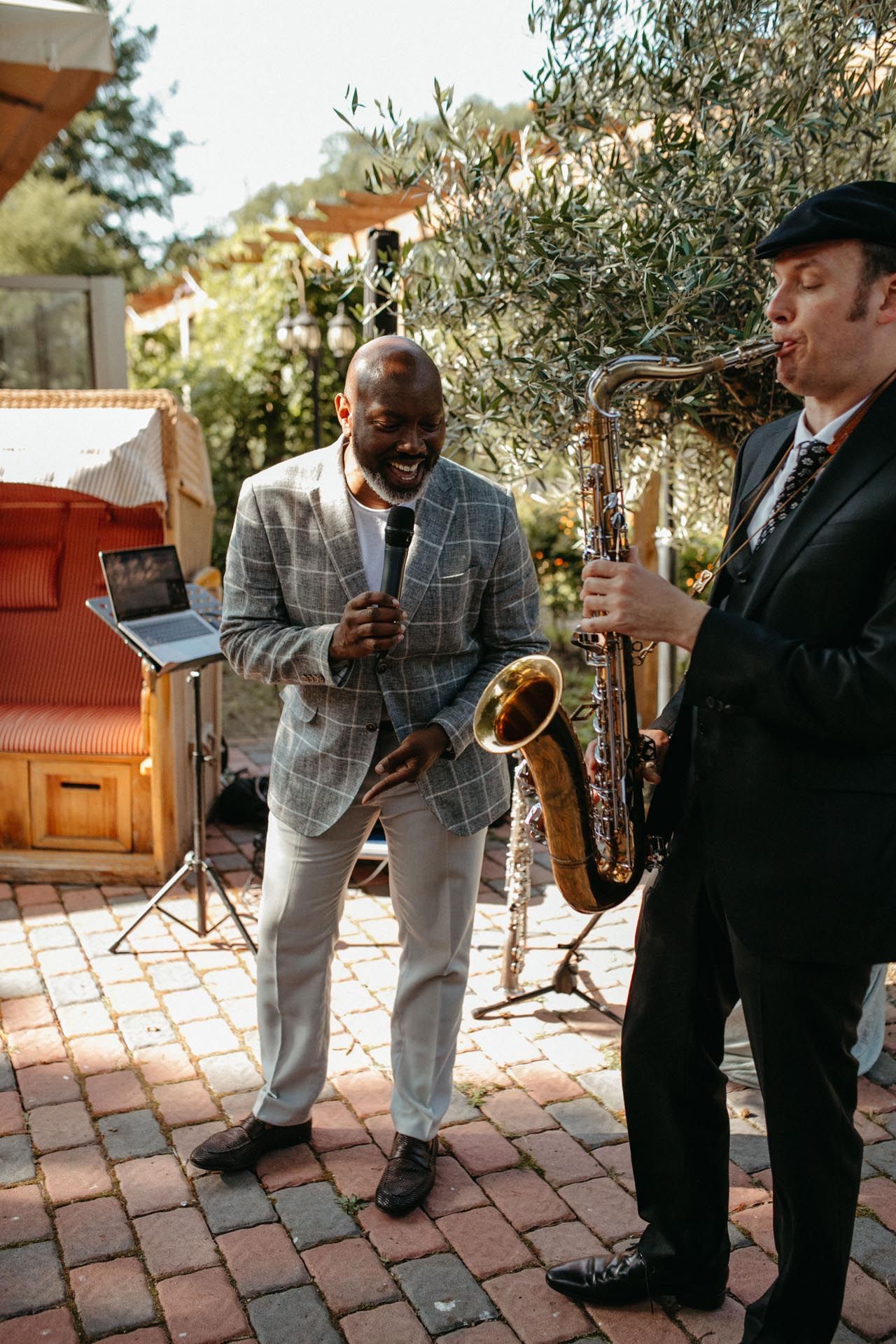 Zwei Musiker treten im Freien auf. Einer singt mit einem Mikrofon, der andere spielt Saxophon. Ziegelterrasse, Grün.