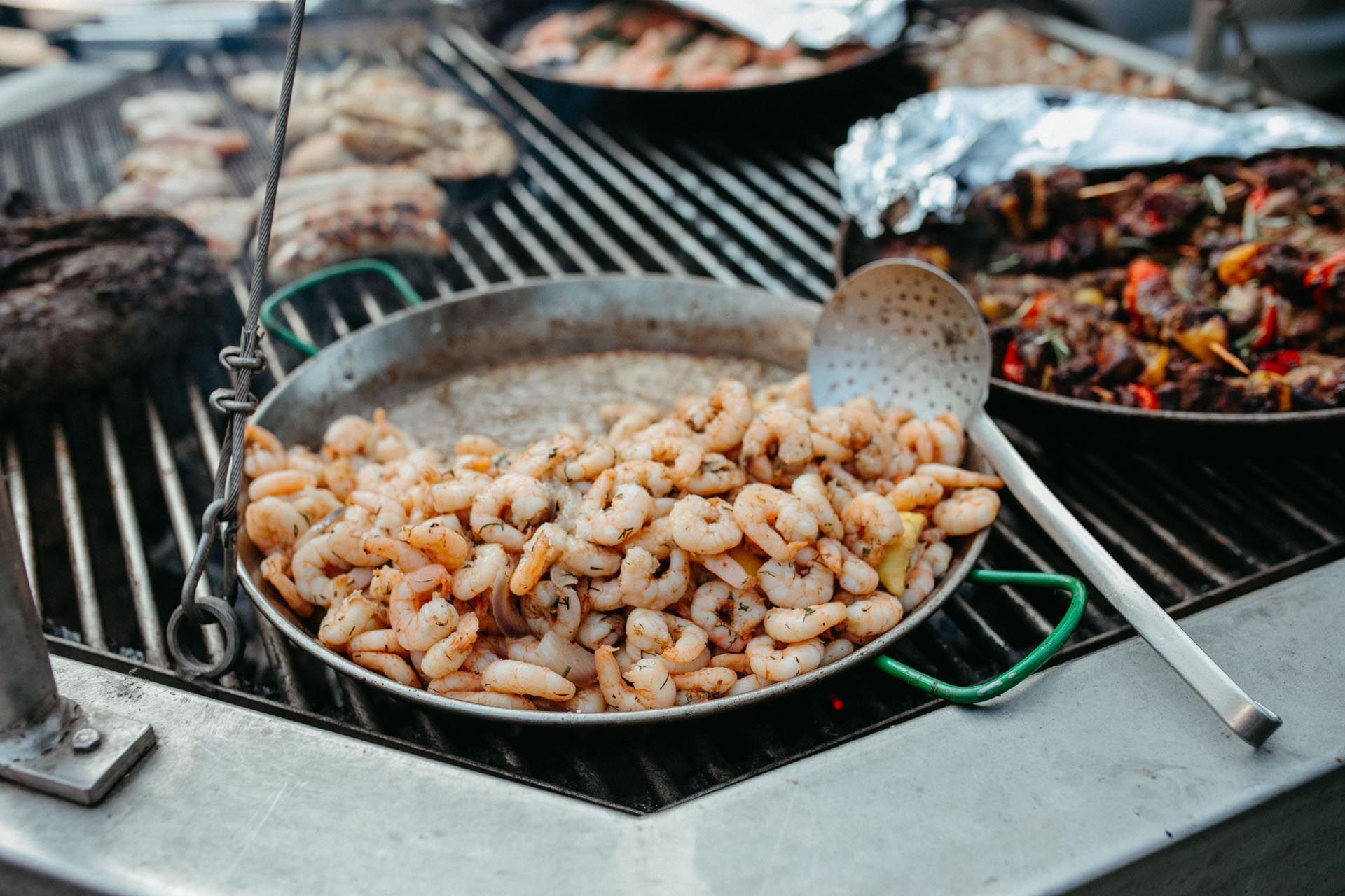Gegrillte Garnelen in einer Pfanne auf einem Grill mit anderen Lebensmitteln, einem Löffel und einer Grilleinstellung.