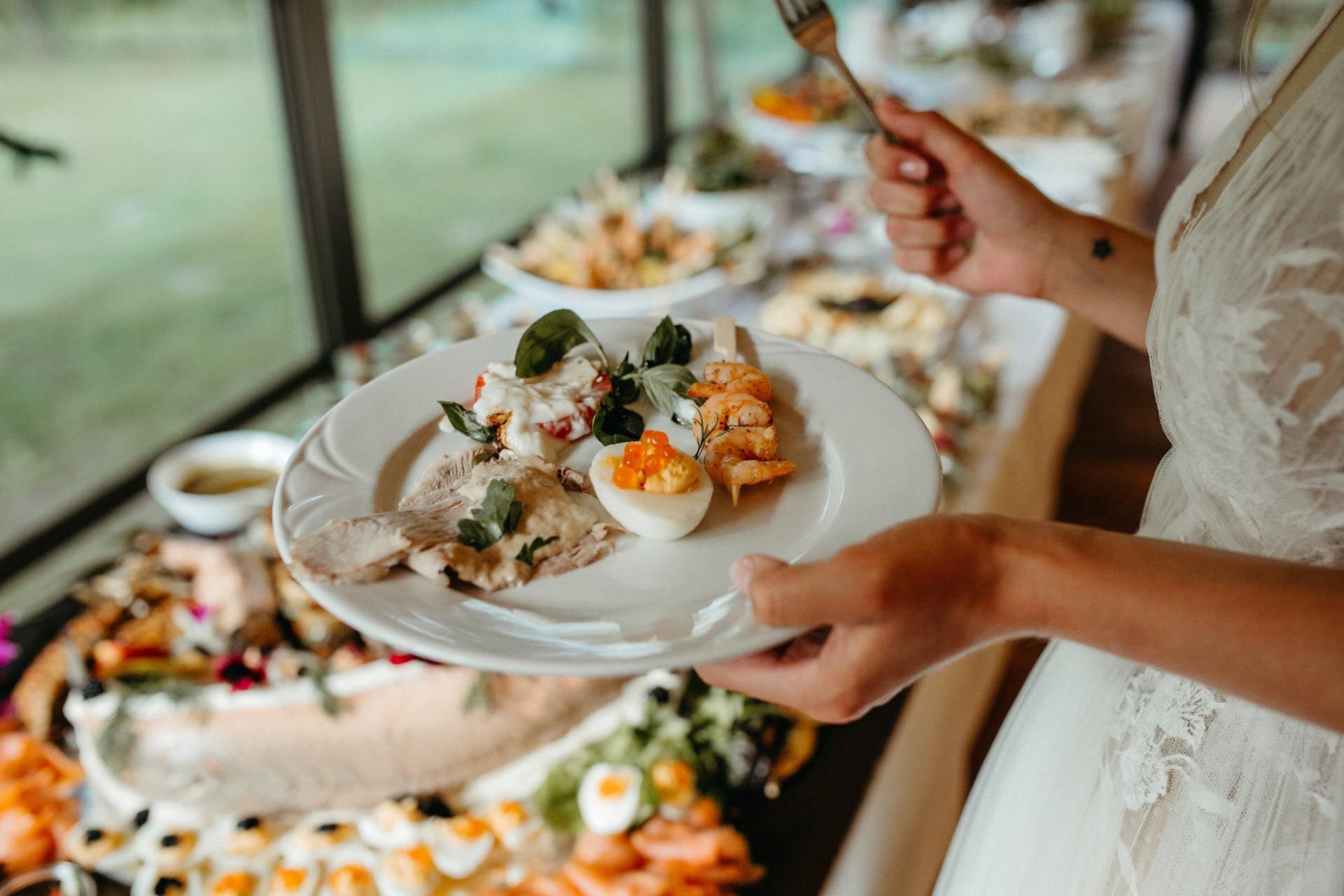 Frau im weißen Kleid hält einen Teller mit Essen neben einem Buffet.