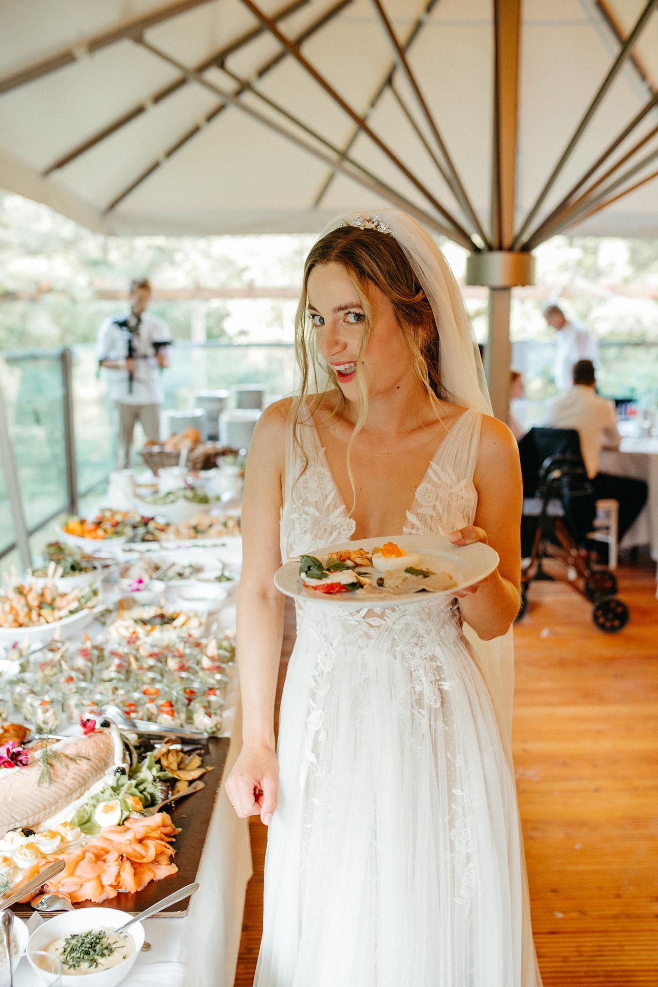 Braut im Brautkleid, die ein Gesicht macht, während sie einen Teller mit Essen an einem Buffet hält.