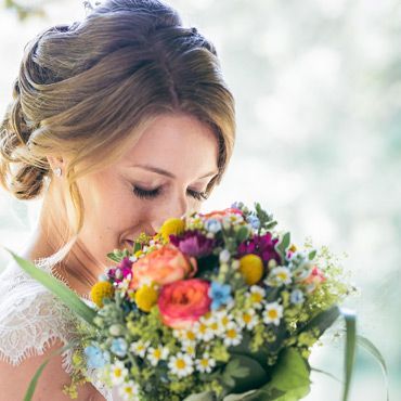 Die Braut riecht an einem bunten Blumenstrauß, wobei der Fokus sanft auf ihrem Gesicht und den Blumen liegt.