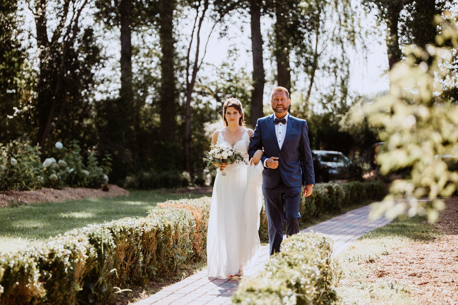 Braut im weißen Kleid geht mit Vater im dunklen Anzug den Weg entlang.