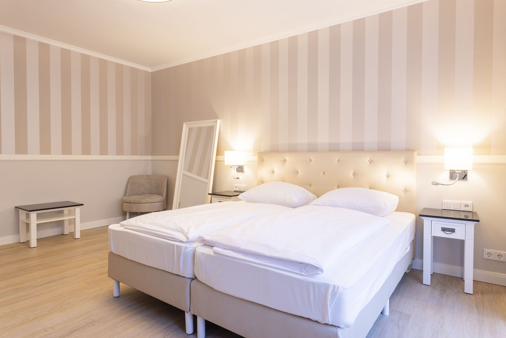 Schlafzimmer mit zwei Betten, hellen Wänden mit Streifen, Stuhl, Spiegel und einem kleinen Tisch.