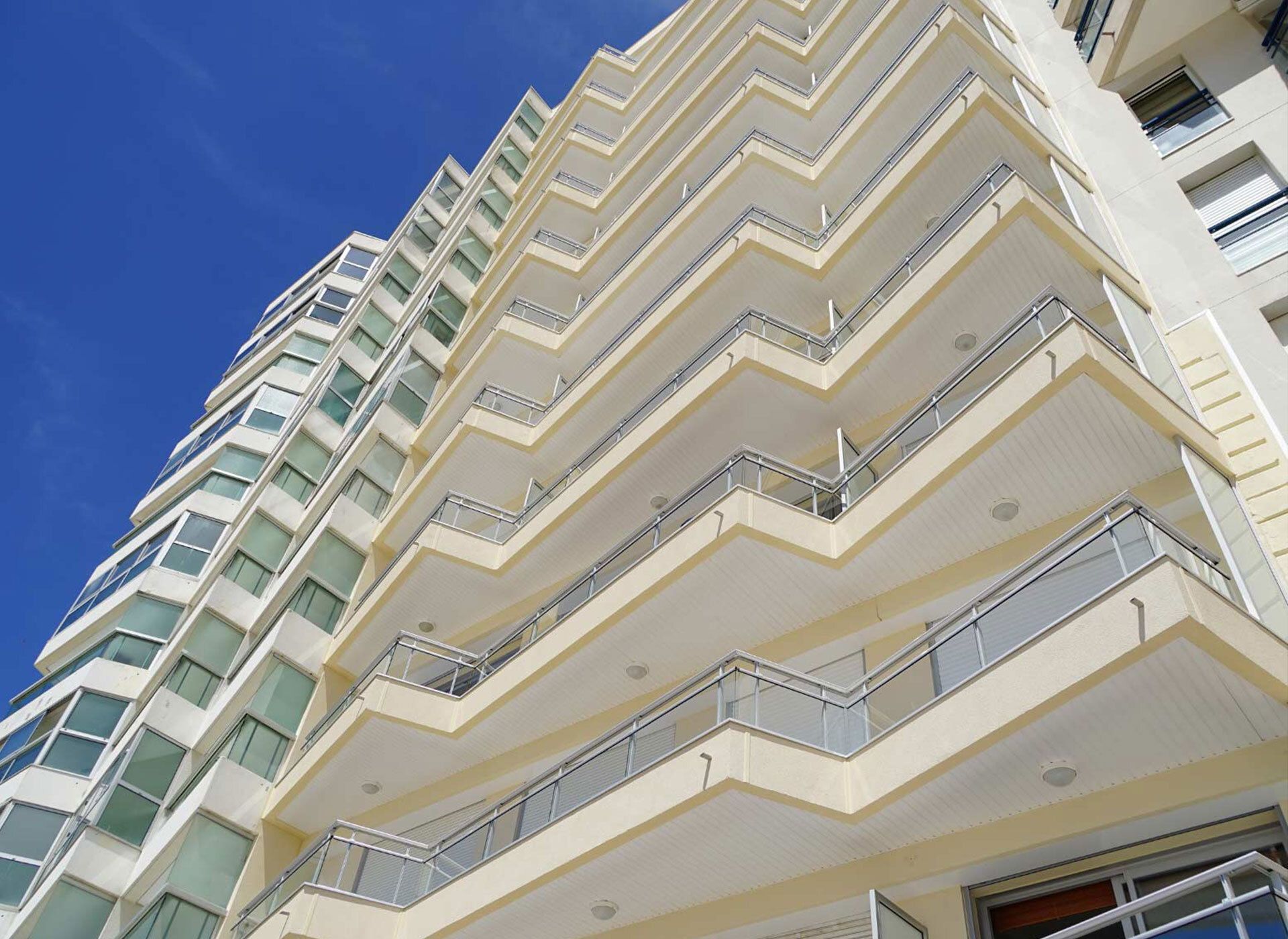 Vue en contre-plongée d'un immeuble moderne à plusieurs étages, doté de balcons en forme de zigzag, se détachant sur un ciel d'un bleu éclatant.