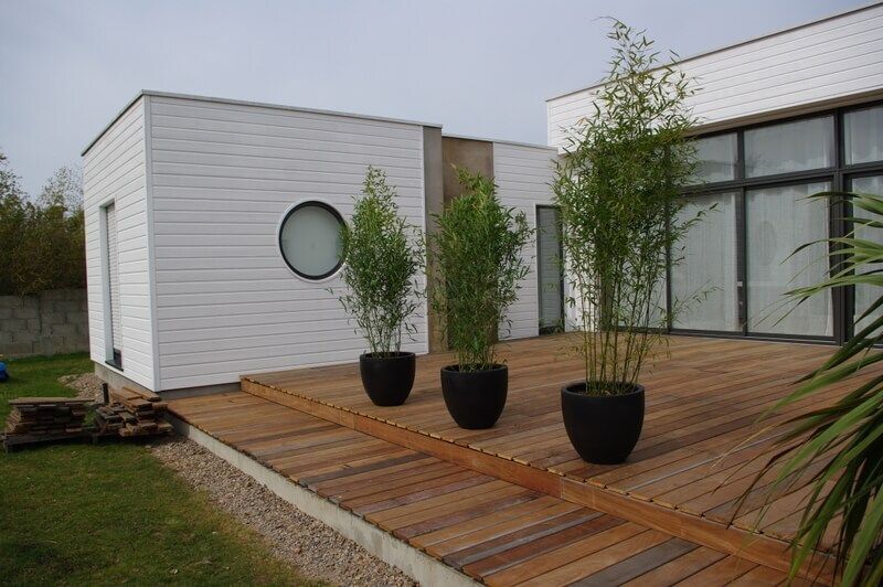 Maison blanche moderne avec terrasse en bois, fenêtre ronde et plantes en pot près de grandes baies vitrées
