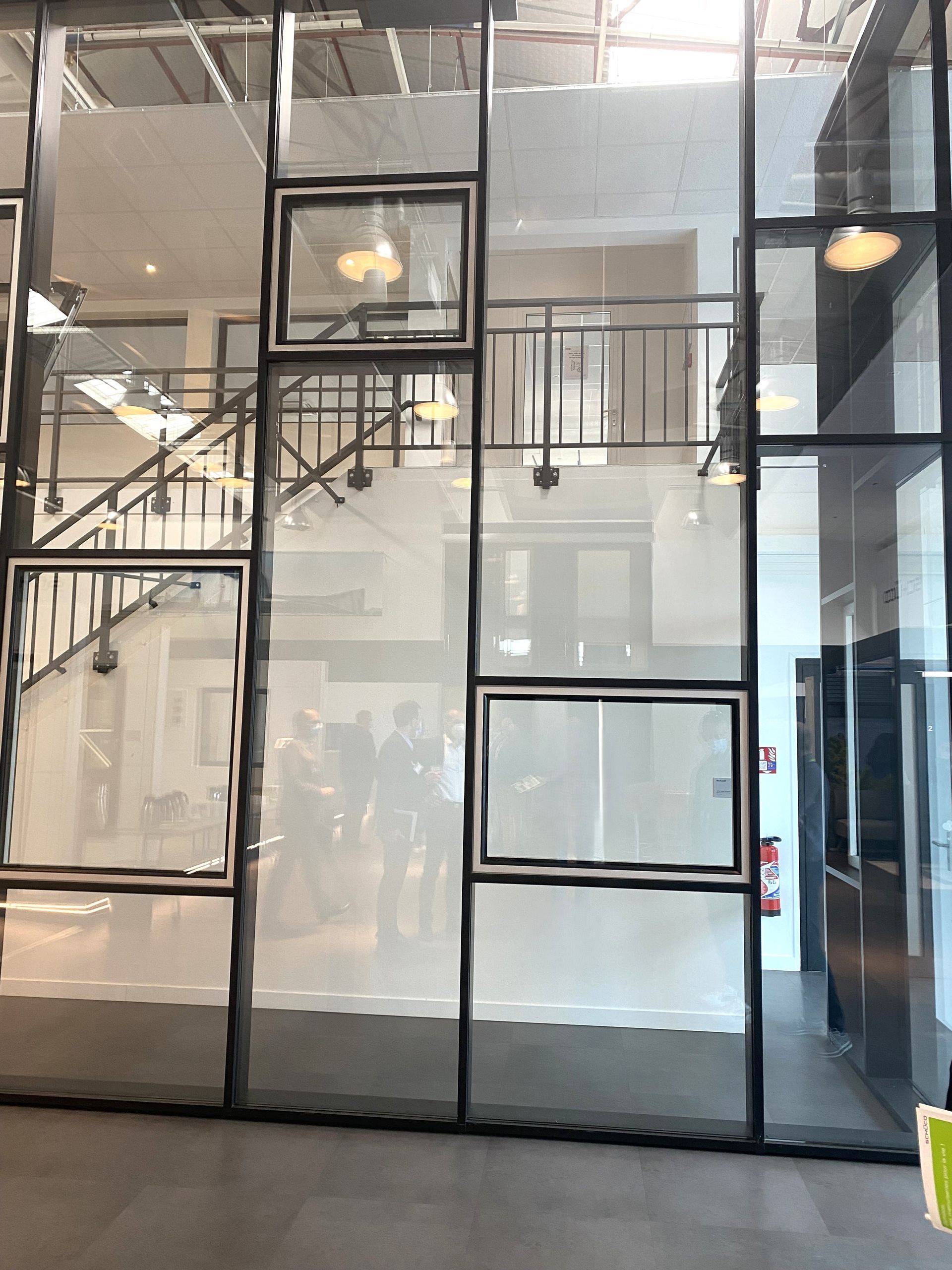 Dans le hall d'un immeuble moderne, une paroi de verre aux cadres métalliques noirs reflète les escaliers et les lumières