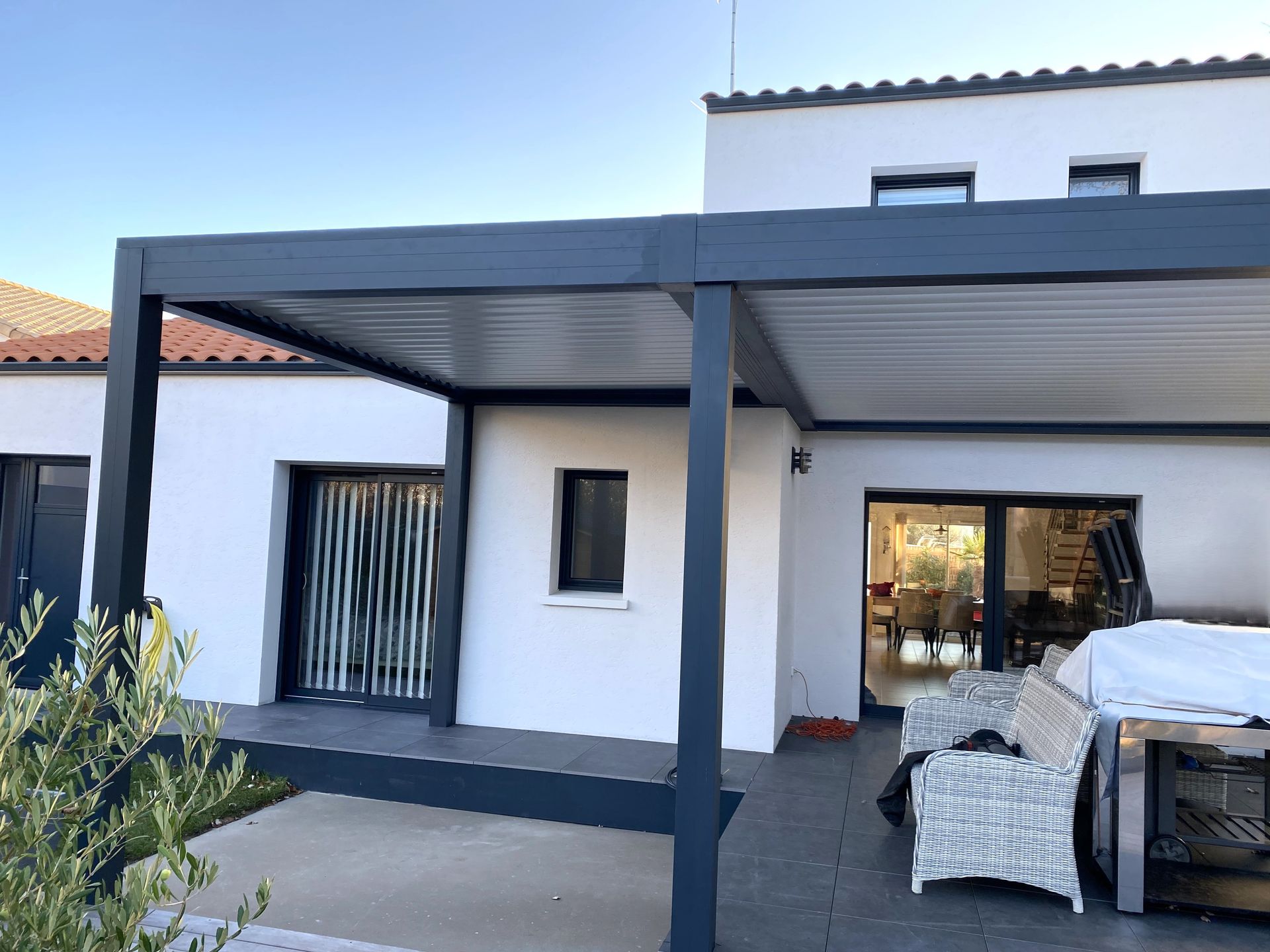 Une pergola gris foncé à toit à lames orientables surplombe un patio jouxtant une maison blanche dotée de portes coulissantes en verre.