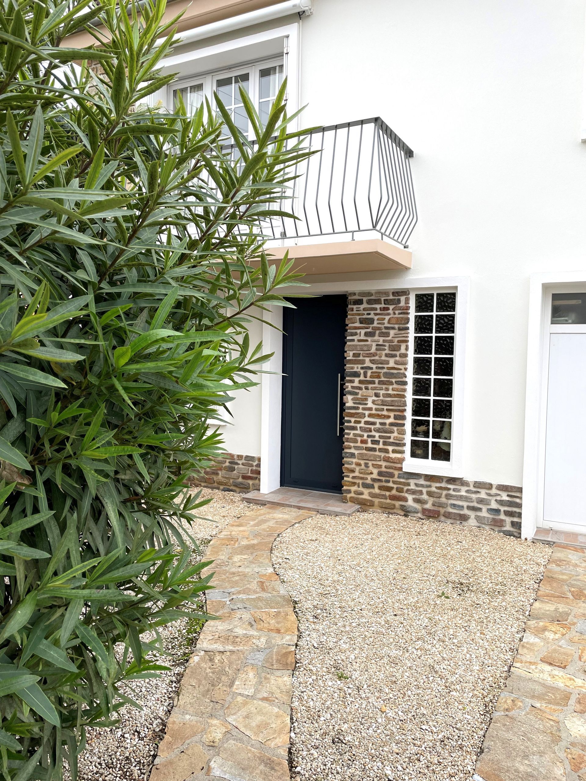 Entrée principale d'une maison blanche avec allée en pierre, cour en gravier et balcon au-dessus de la porte