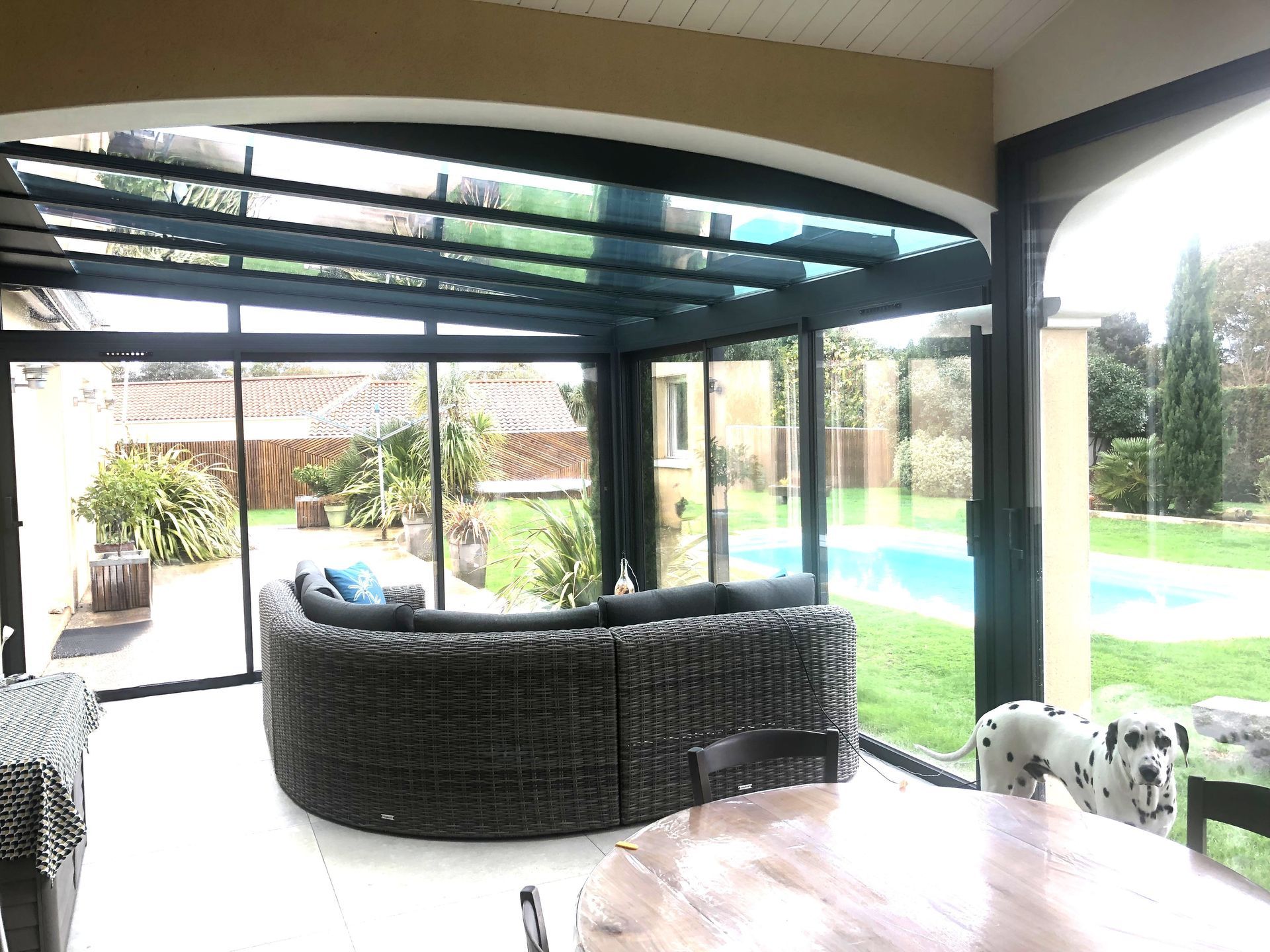 Terrasse couverte avec sièges en osier, toit en verre et vue sur la piscine et le jardin