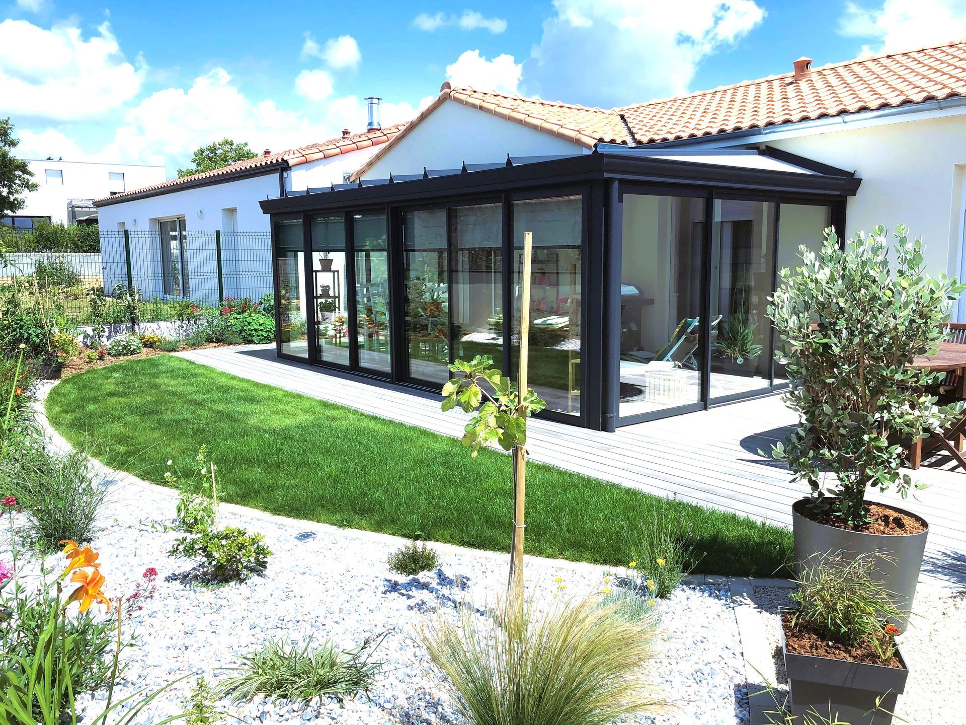 Maison moderne avec véranda vitrée, jardin de gravier blanc et plantes en pot sous un ciel bleu