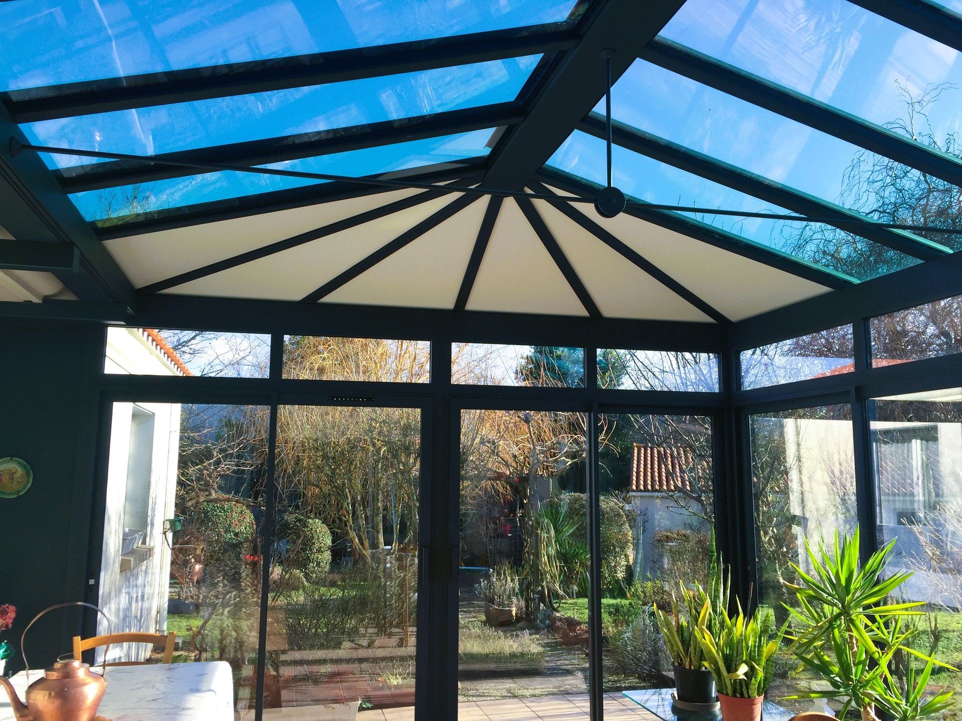 Une pergola moderne en métal gris foncé, adossée à une maison blanche, avec une terrasse et du mobilier de jardin.