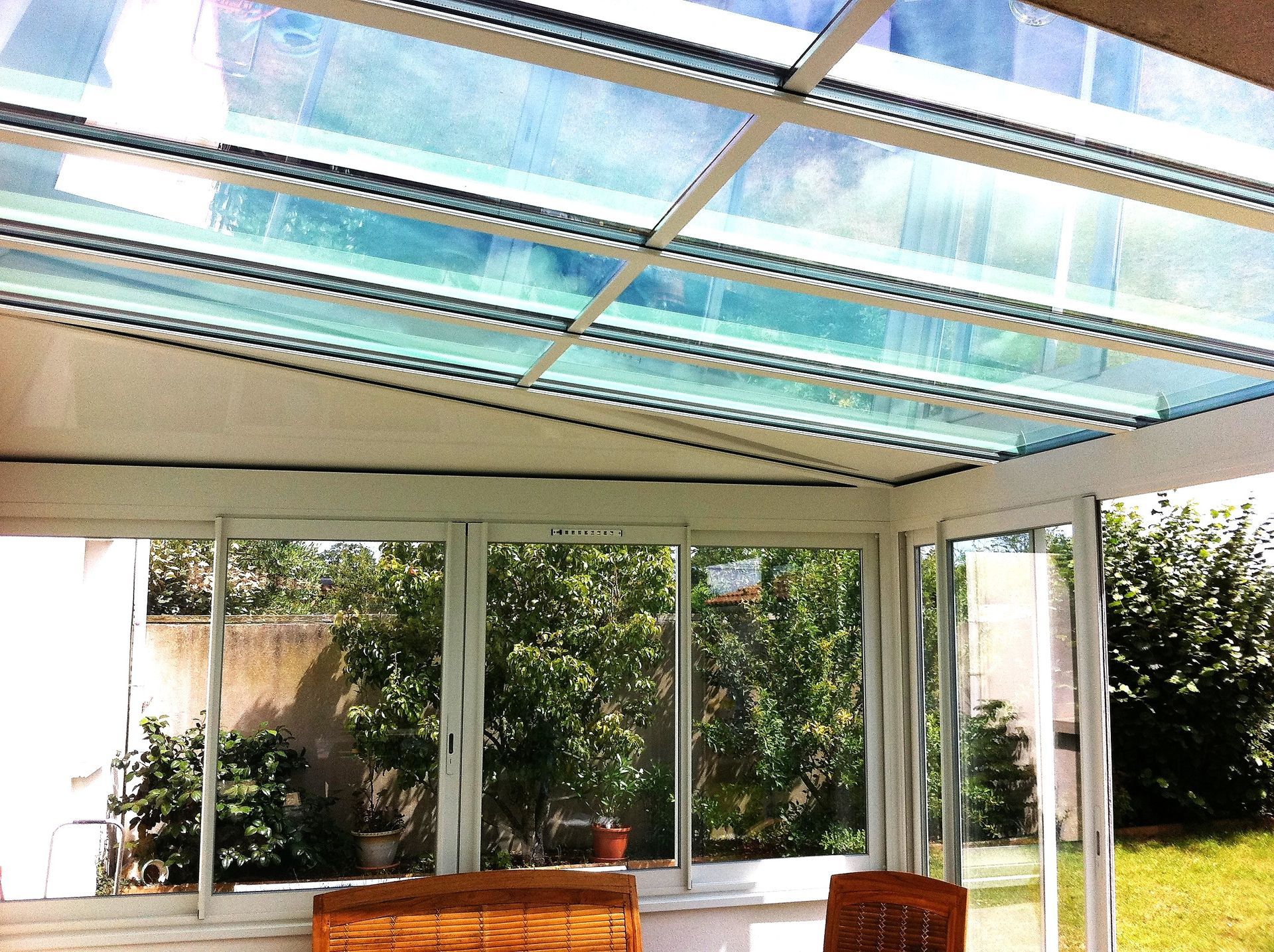 Patio ensoleillé avec toit en verre, grandes fenêtres et banc en bois donnant sur un jardin verdoyant