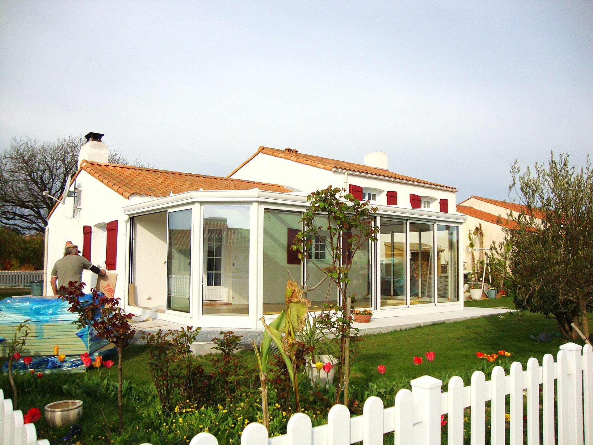 Maison blanche au toit rouge, véranda vitrée et clôture blanche dans un jardin ensoleillé