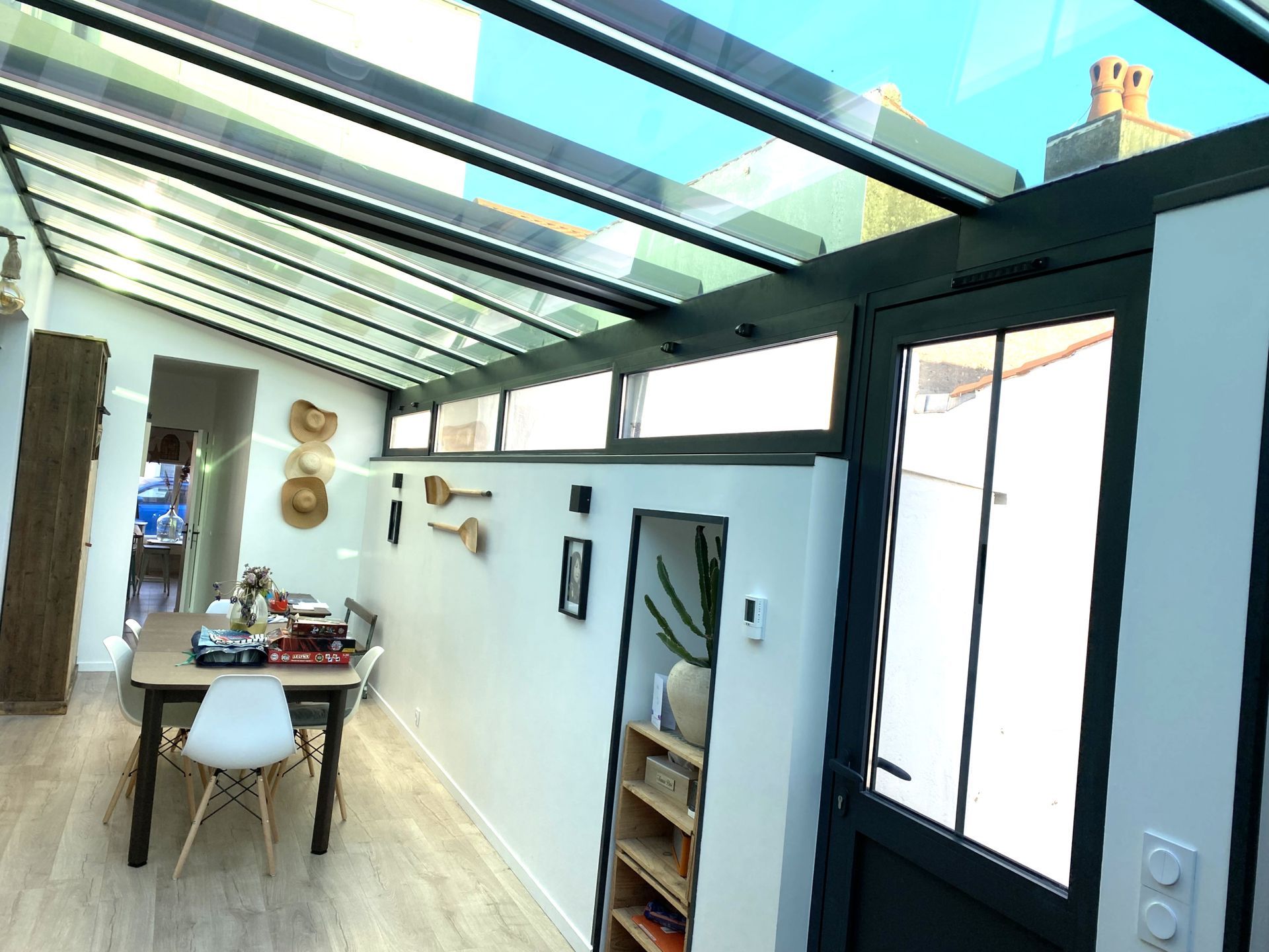 Véranda lumineuse avec toit en verre, longue table à manger, chaises blanches et un couloir étroit sur un côté