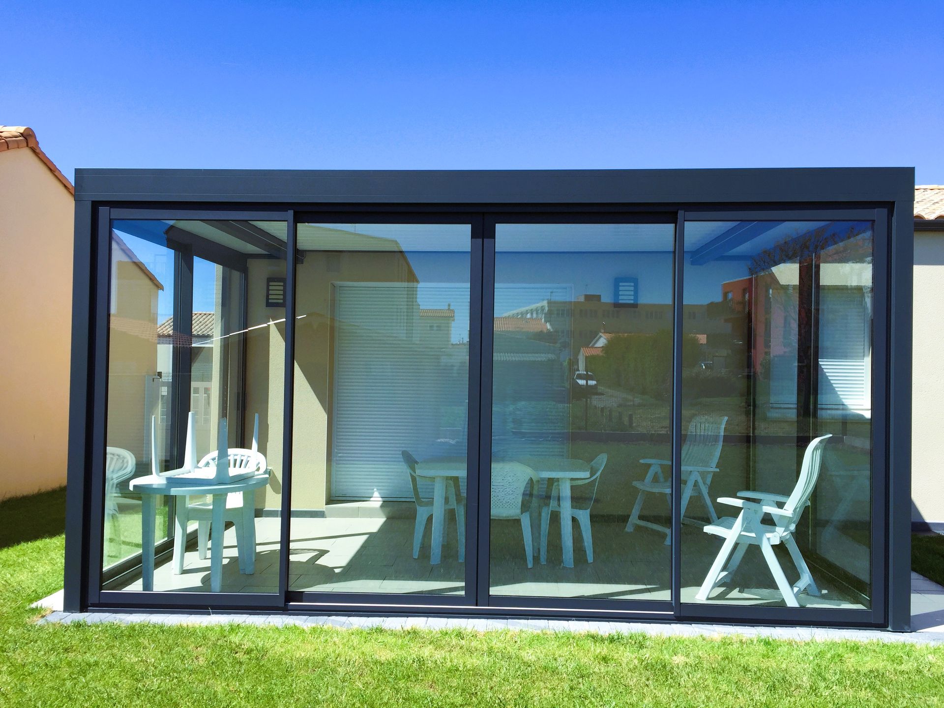 Patio moderne aux parois de verre, avec chaises et table blanches, reflétant une scène extérieure ensoleillée