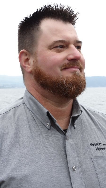 Mann mit rotem Bart und grauem Hemd blickt zur Seite. Im Hintergrund verschwimmen Wasser und Himmel.