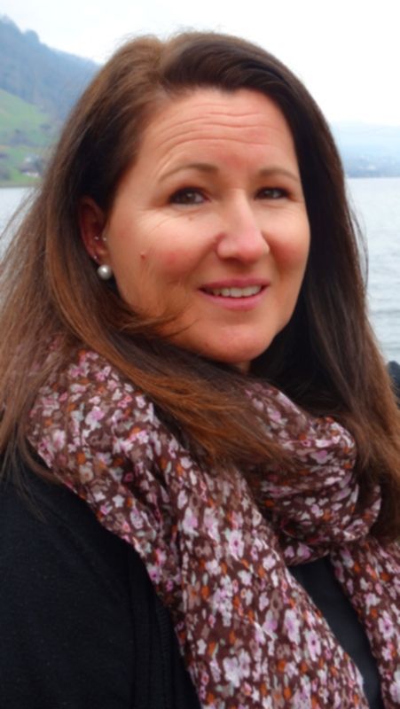 Frau mit braunem Haar, lächelnd, mit einem geblümten Schal, vor einer Kulisse aus See und Bergen.