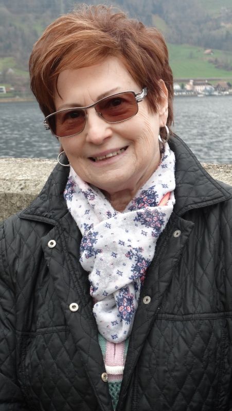 Frau mit Sonnenbrille und Steppjacke lächelt im Freien, Schal, See im Hintergrund.