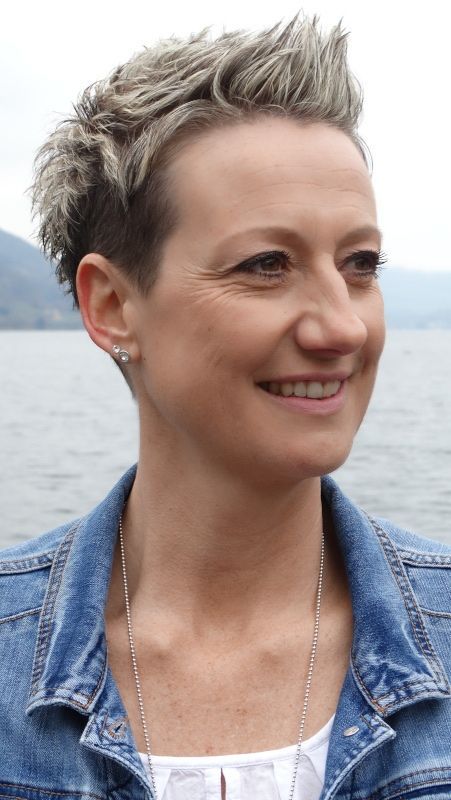 Frau mit kurzen, stacheligen blonden Haaren, lächelnd, trägt eine Jeansjacke, im Freien in der Nähe von Wasser.