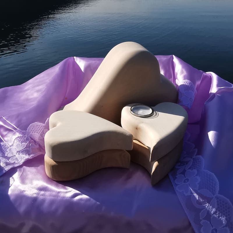 Drei herzförmige beige Urnen auf einem lila Satintuch mit Spitzenbesatz; im Hintergrund ein Gewässer.