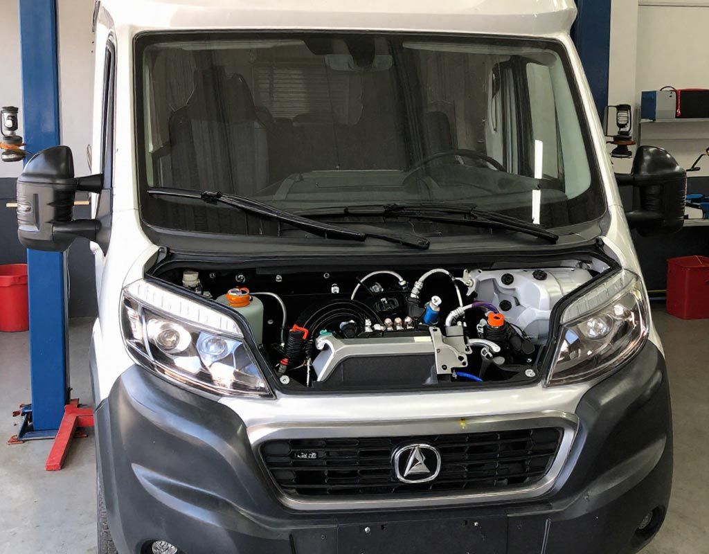 Una furgoneta de reparto blanca aparcada en un garaje cubierto con el capó abierto, dejando al descubierto los componentes del motor.