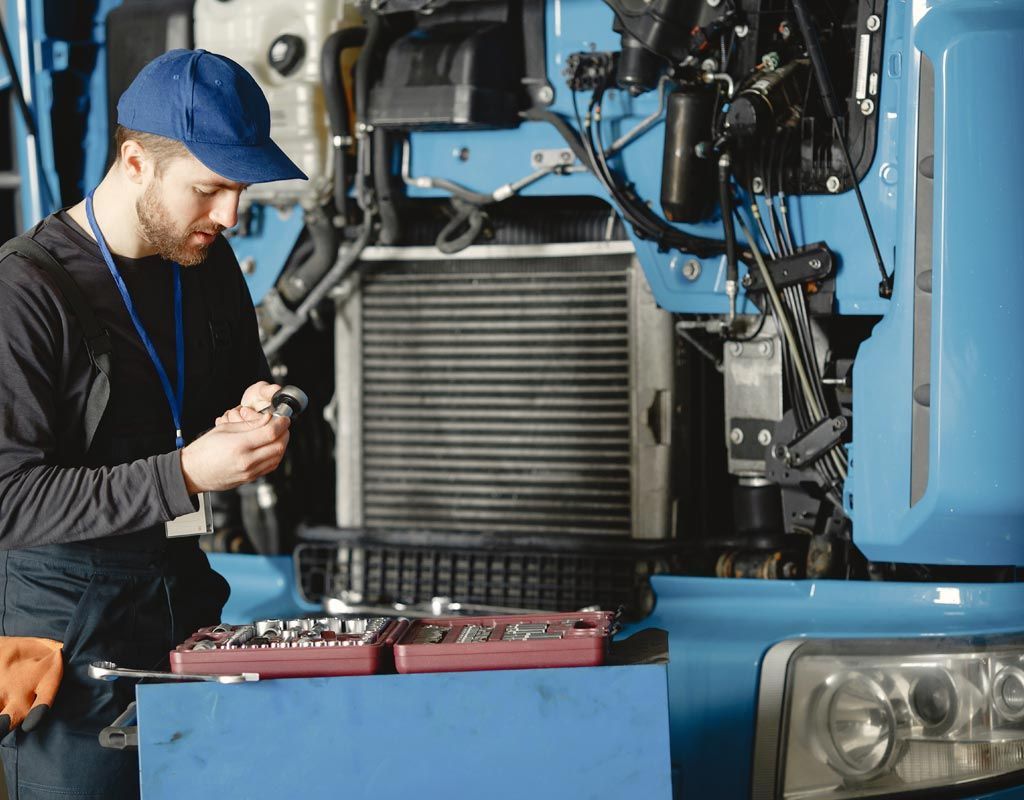 Un técnico con gorra azul y uniforme de trabajo utiliza una herramienta mientras repara el motor de un camión azul.
