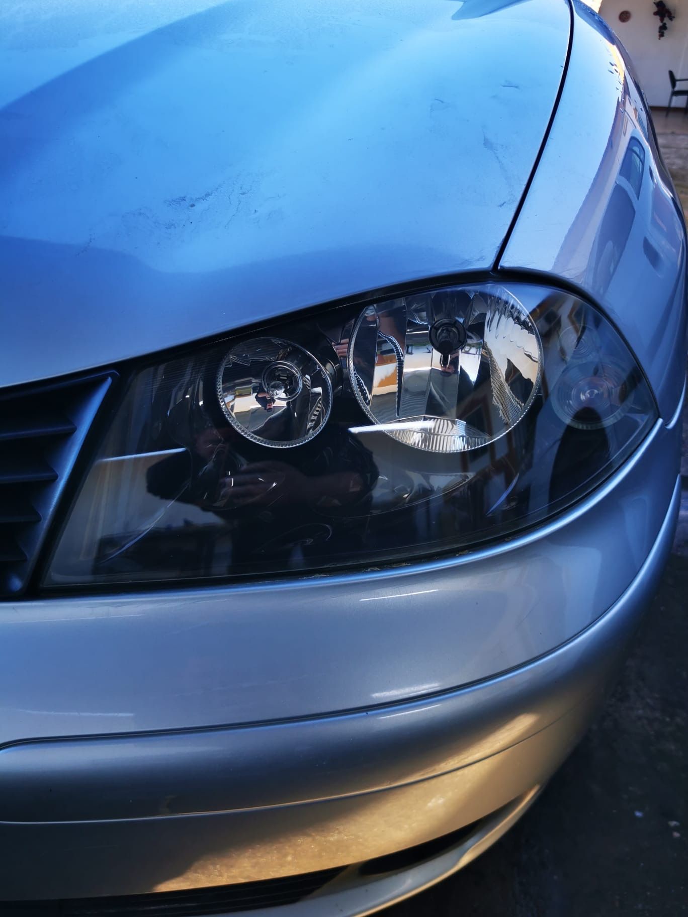 Parte frontal de un coche plateado con faros tintados que reflejan el entorno.