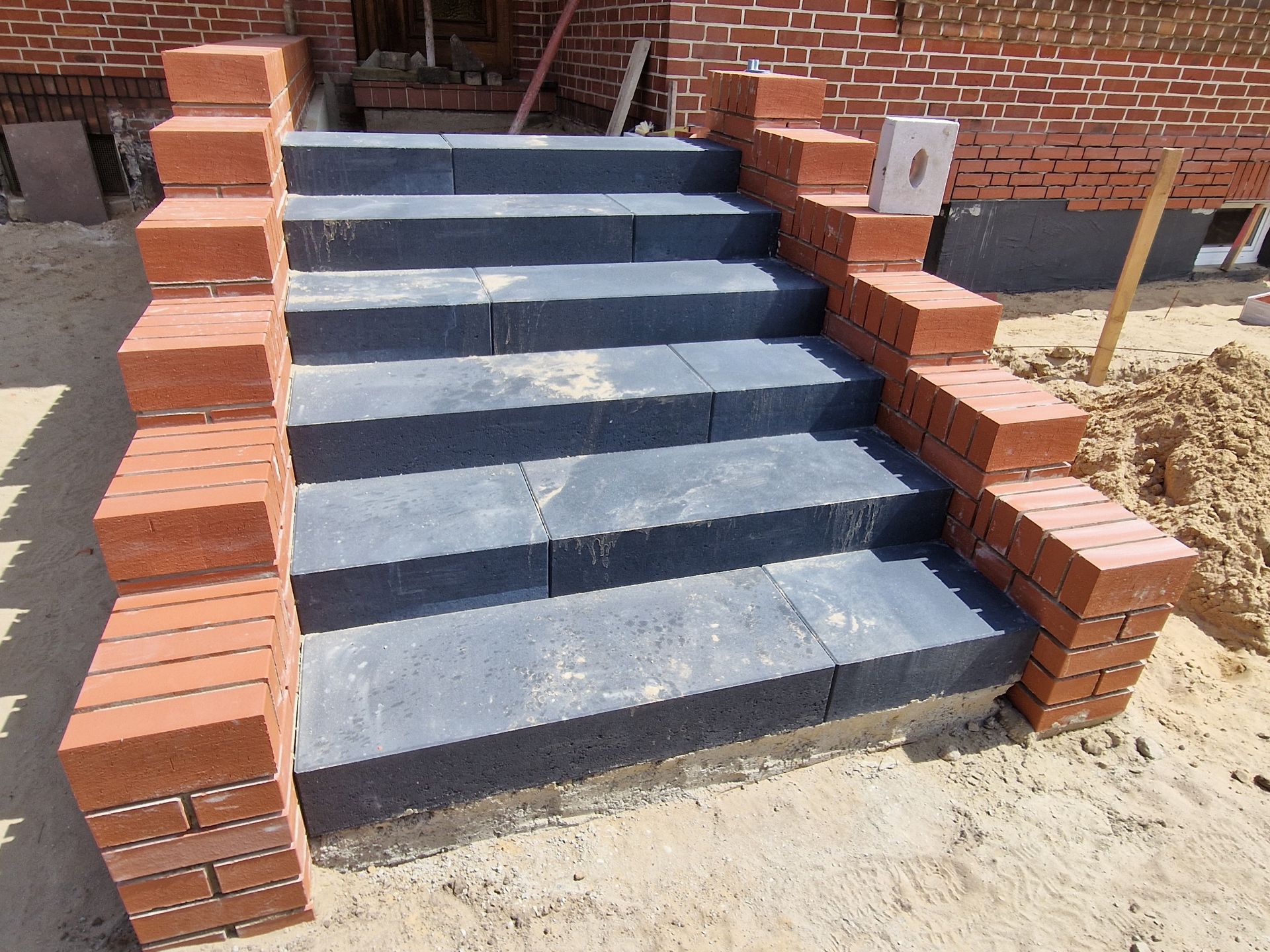 Neu errichtete Treppe mit dunklen Steinstufen, flankiert von roten Backsteinsäulen, in einem sandigen Baugebiet.