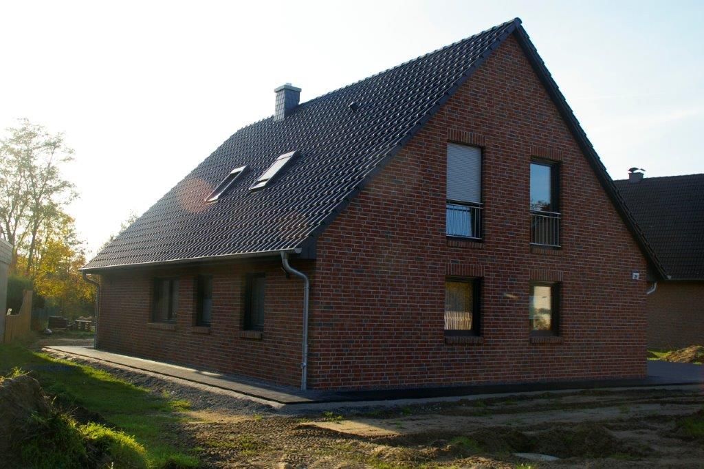 Ein modernes Backsteinhaus mit schrägem Dach, mehreren Fenstern und einem kleinen Garten im Vordergrund, bei klarem Himmel.