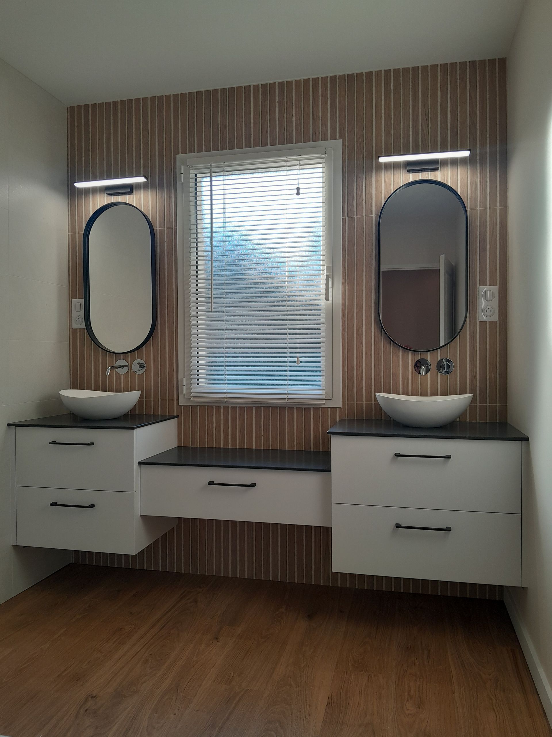 Salle de bains moderne avec vanités flottantes, miroirs ovales et carrelage imitation bois.