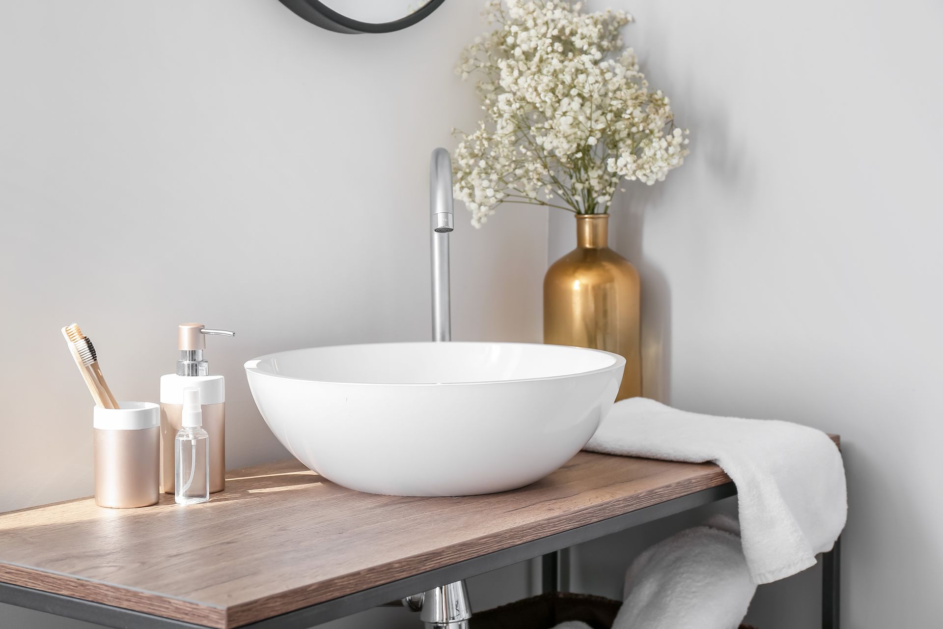 Lavabo blanc sur un comptoir en bois avec des articles de toilette, un vase de fleurs doré et une serviette dans une salle de bains.
