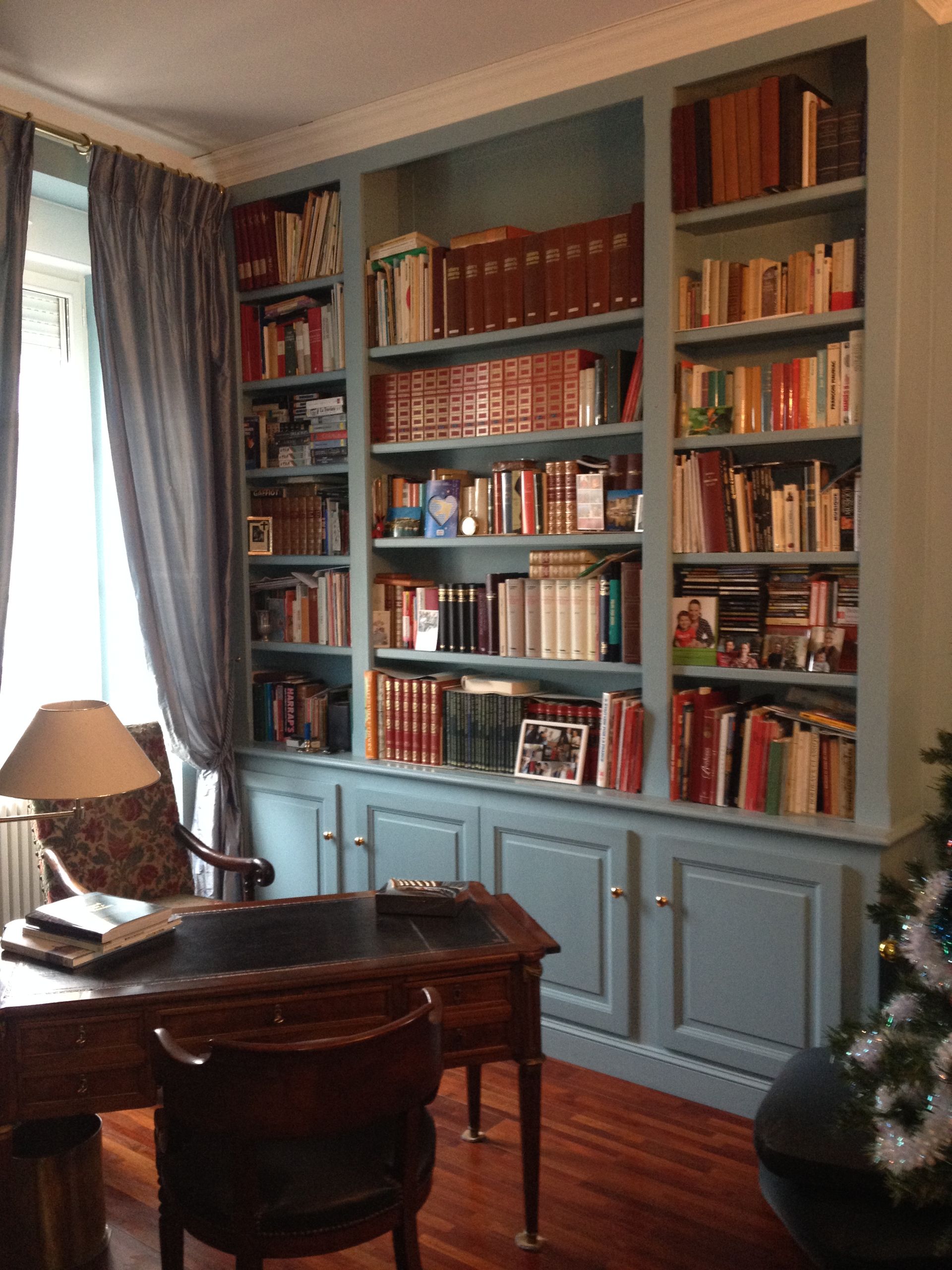 Étagères bleues intégrées remplies de livres, au-dessus d'un bureau et d'une chaise dans une pièce avec un rideau.