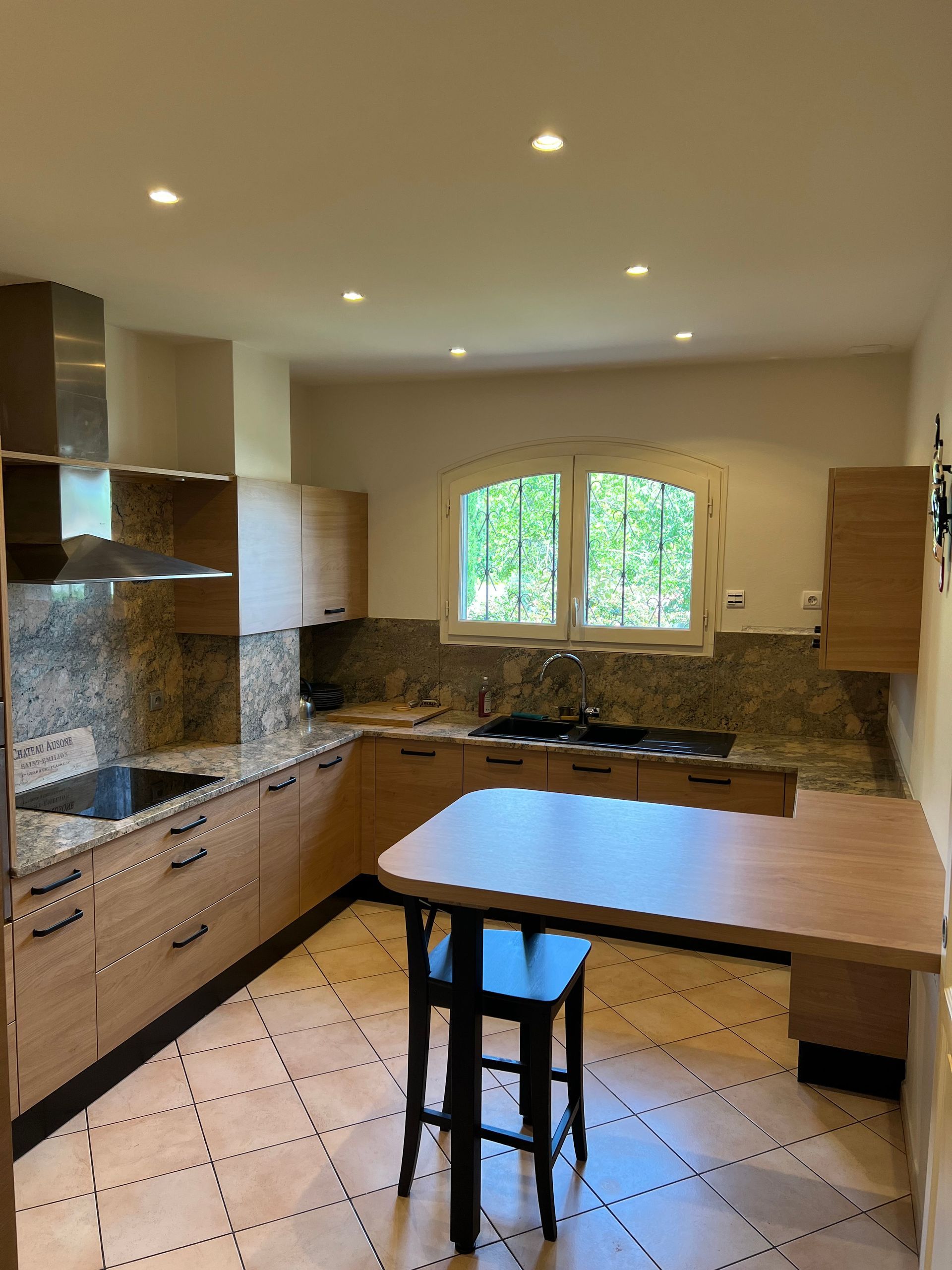 Cuisine avec armoires en bois, comptoirs en granit, îlot avec tabouret noir et lumière naturelle provenant de la fenêtre.