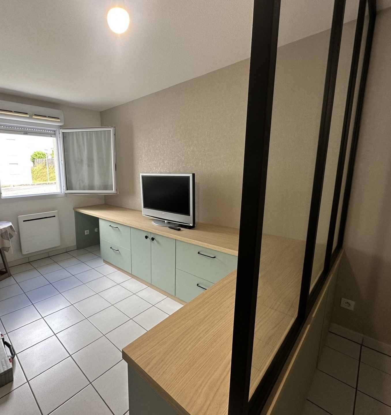 Une pièce avec un téléviseur posé sur un meuble vert menthe et un comptoir en bois. Des poutres métalliques noires créent une séparation partielle.