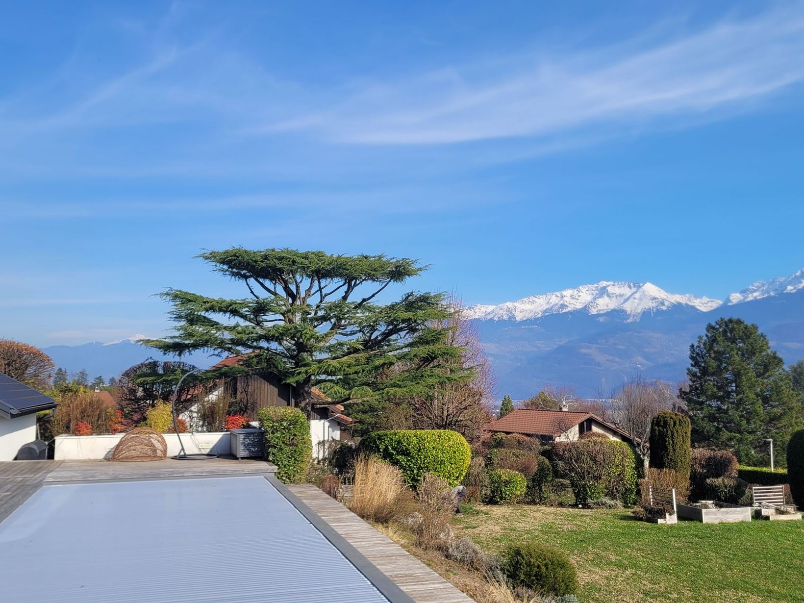 Route de montagne bordée d'arbres et de maisons, avec des sommets enneigés sous un ciel d'un bleu limpide.