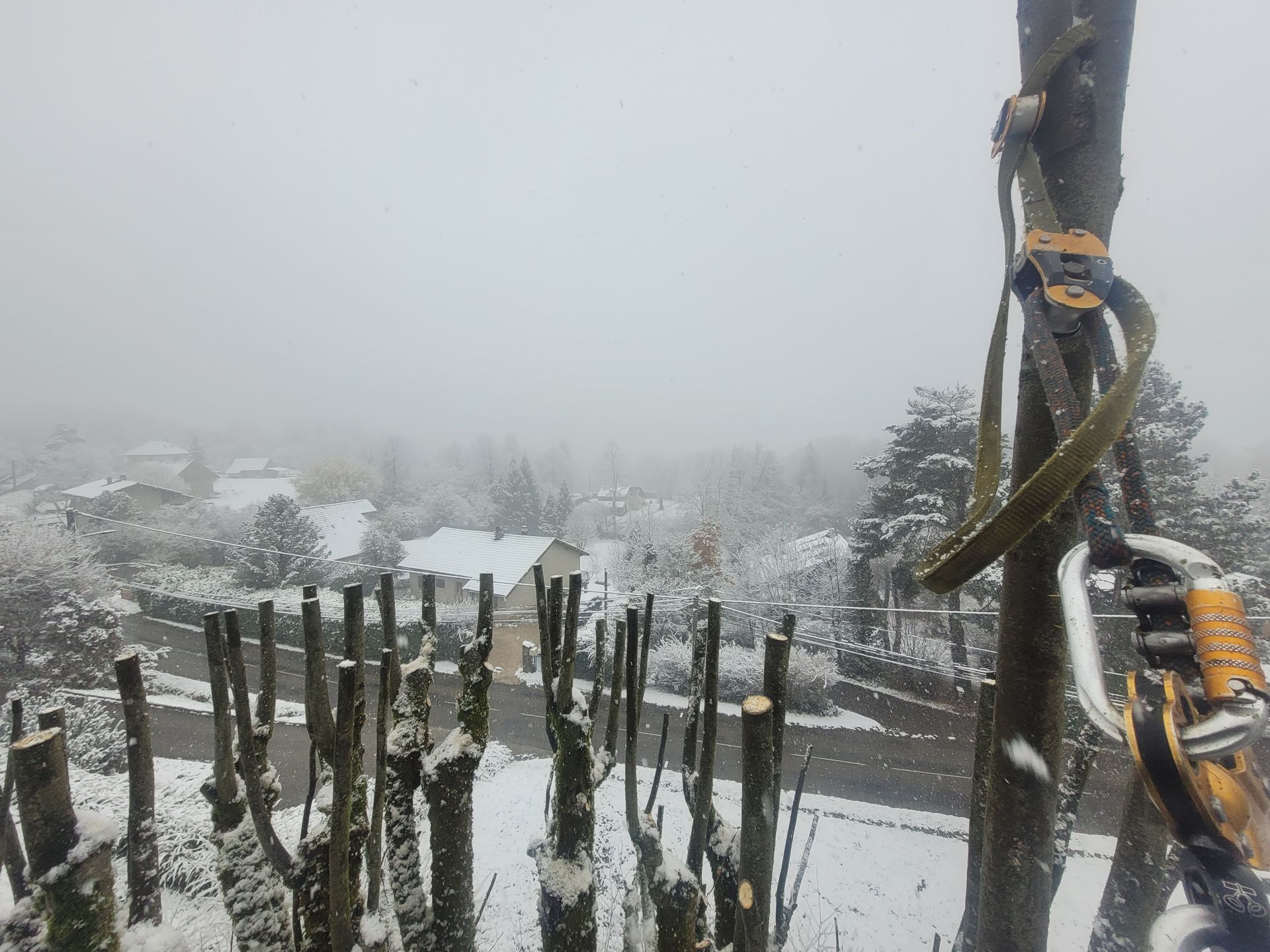 Village enneigé et brumeux à flanc de colline, vu d'un balcon en bois, avec un poteau recouvert de corde au premier plan.