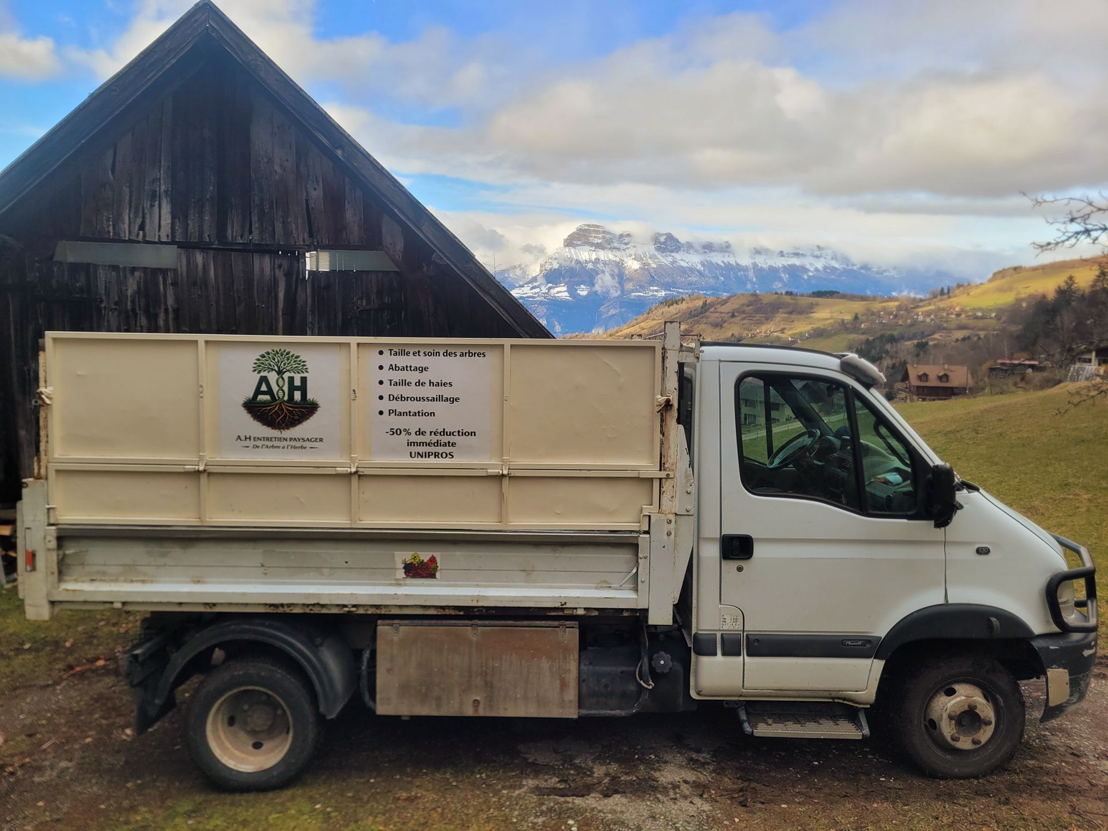 Un camion-benne blanc est garé près d'une grange rustique, avec des montagnes enneigées en arrière-plan.
