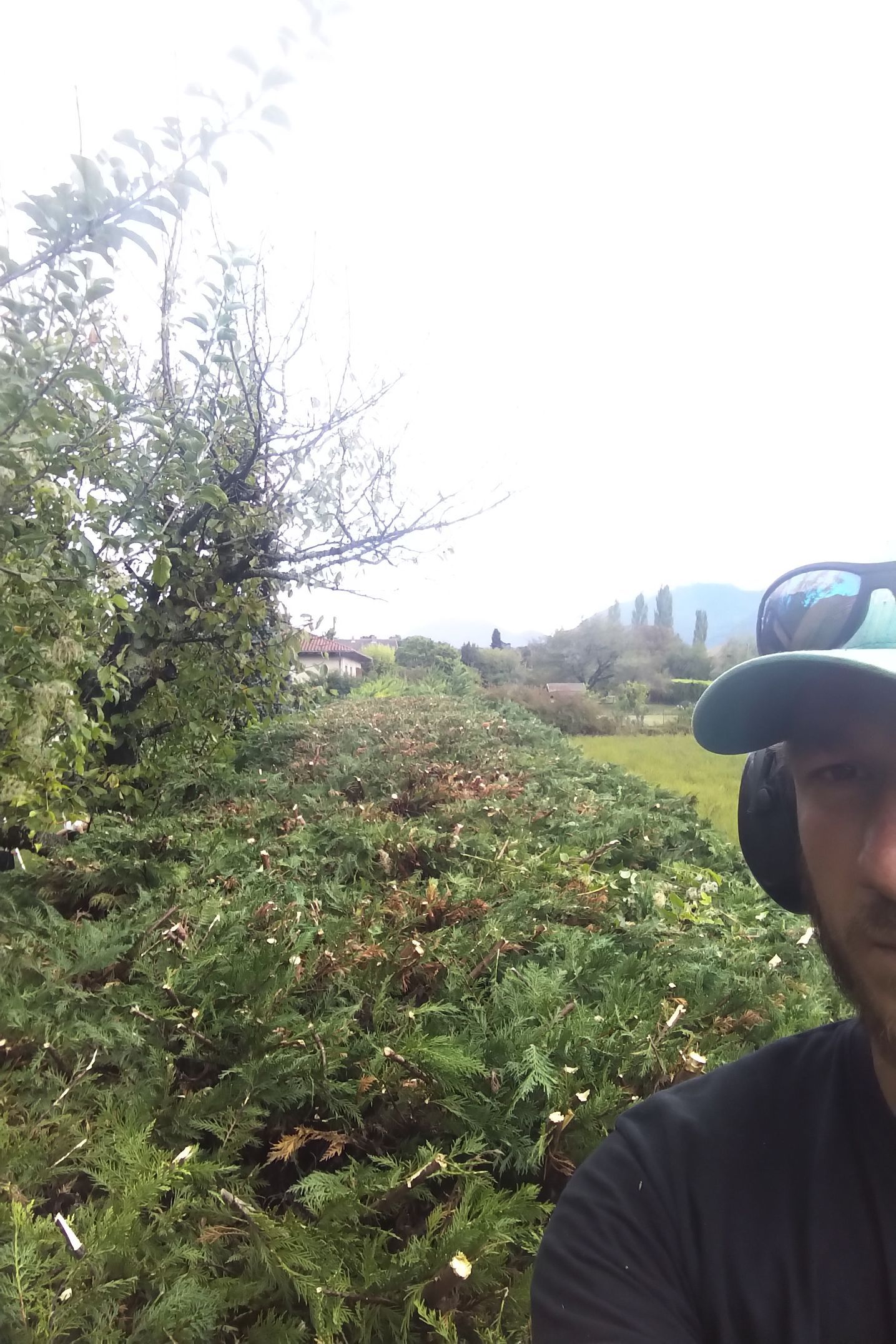 Une personne prend un selfie sur un sentier à flanc de colline, bordé d'arbustes verts denses et sous un ciel nuageux.