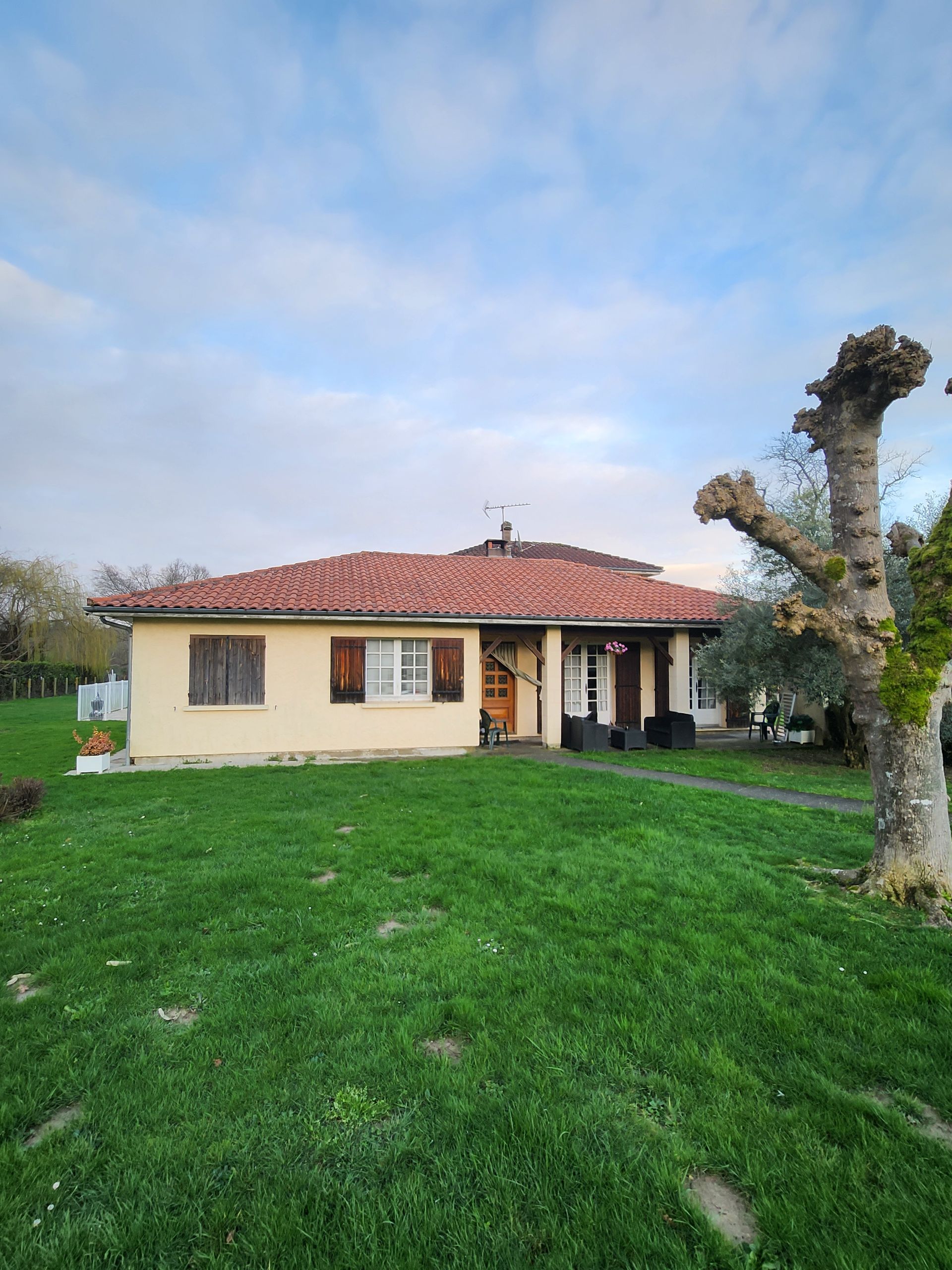 Une maison beige de plain-pied avec un toit de tuiles rouges et un porche couvert, située sur une pelouse verte avec un grand arbre taillé à proximité.