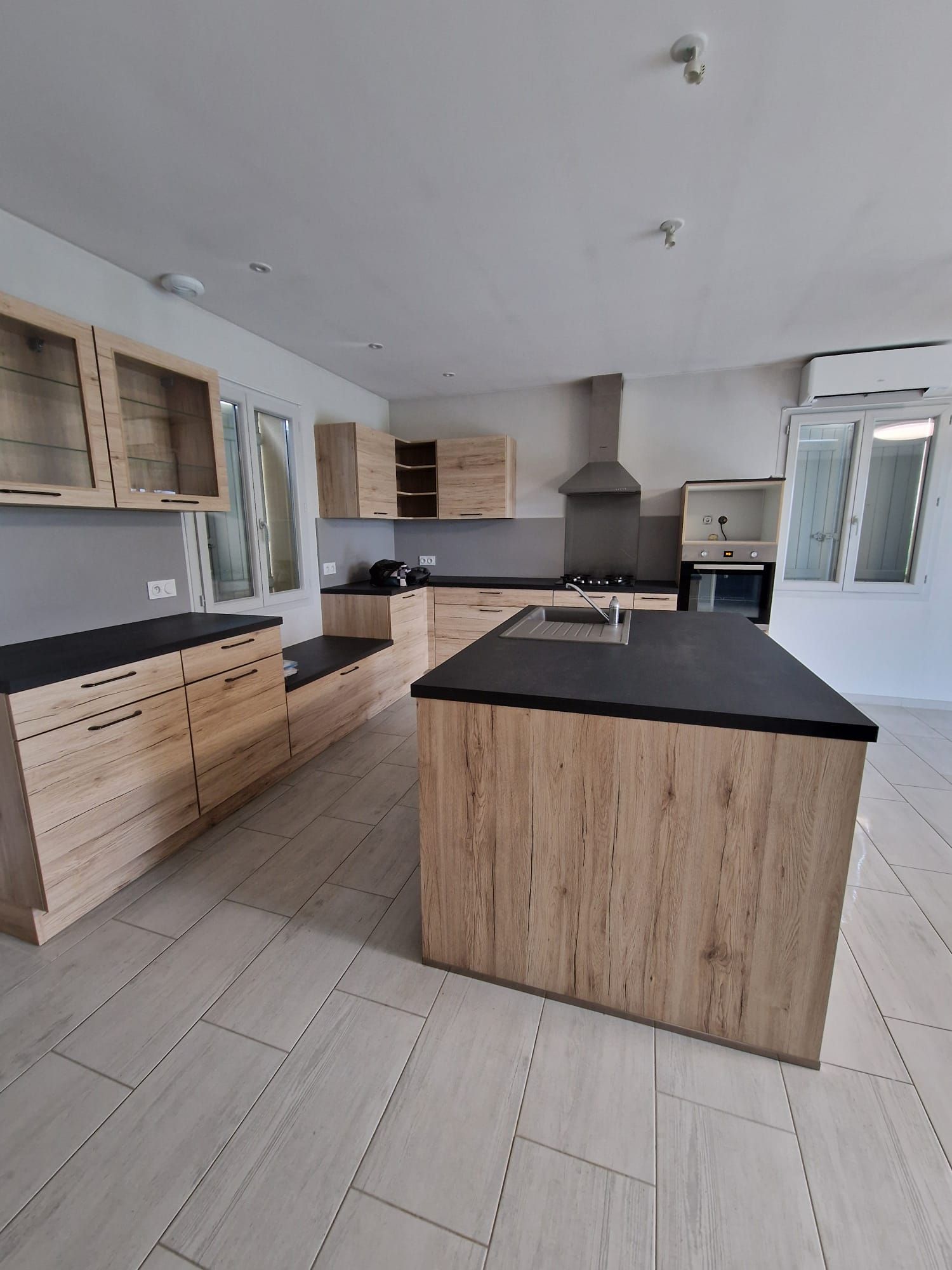 Une cuisine moderne avec des armoires en bois clair, des comptoirs noirs, un îlot central et un sol carrelé.