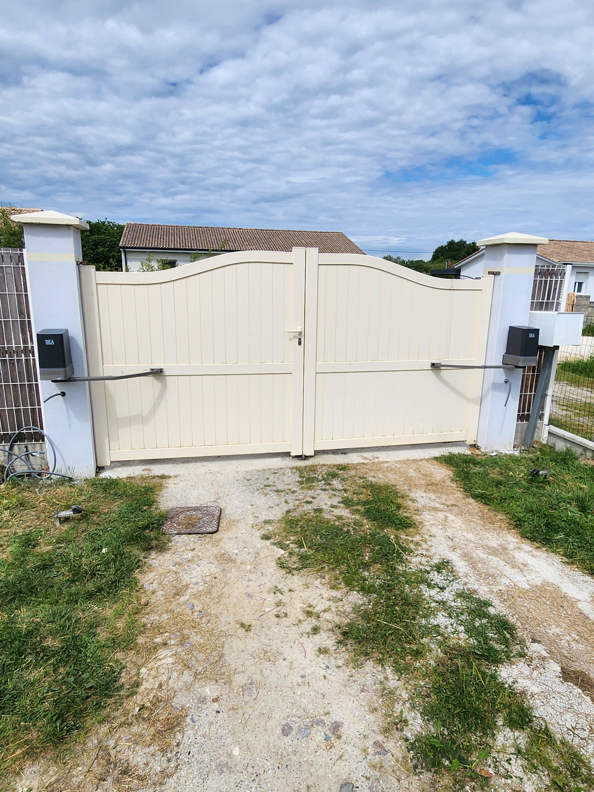 Portail double couleur crème à sommet arqué, monté sur des piliers en pierre avec des bras d'ouverture mécaniques, sur une allée de gravier.