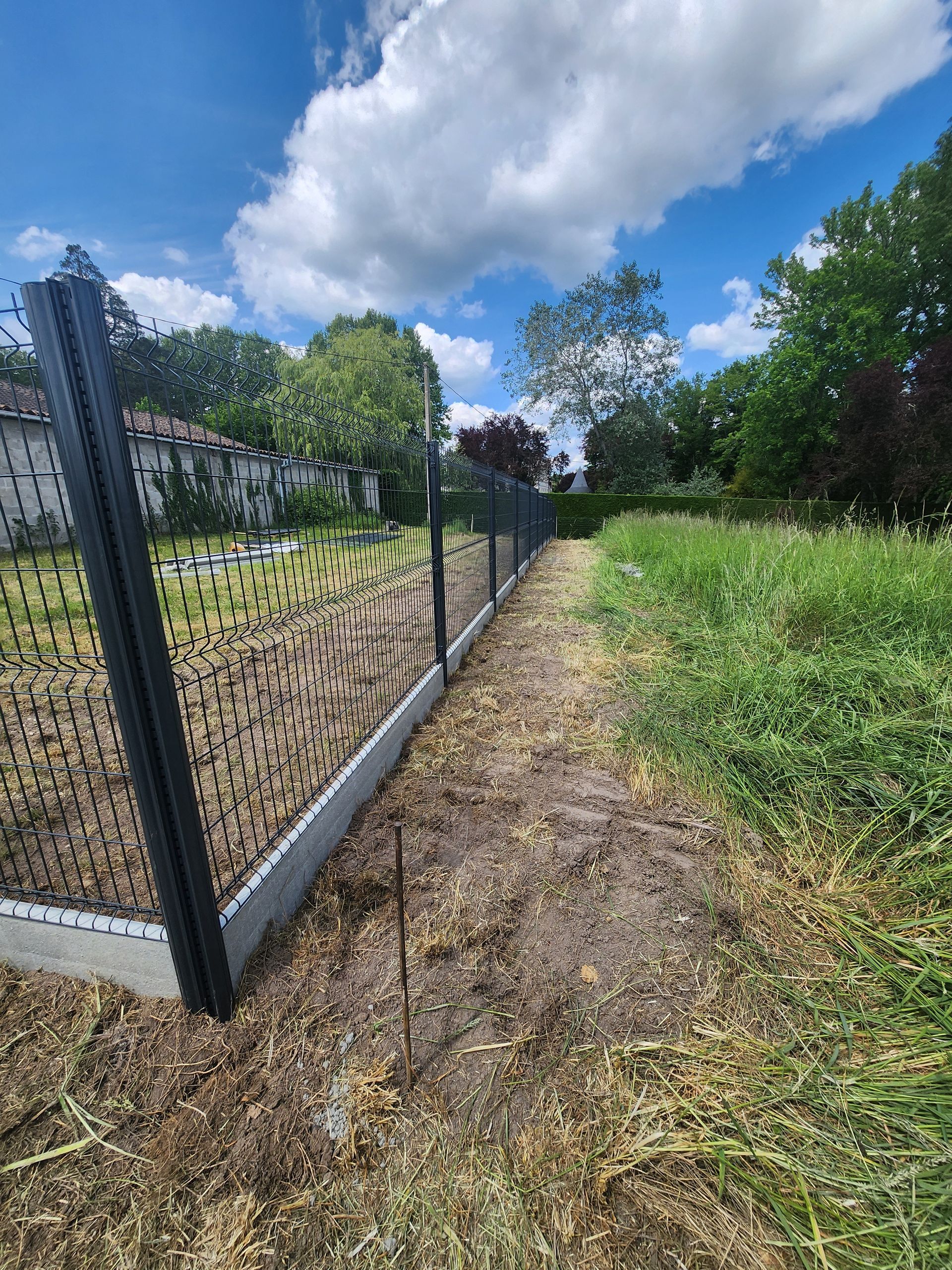 Une clôture en fil de fer noir sur socle en béton longe un champ herbeux.