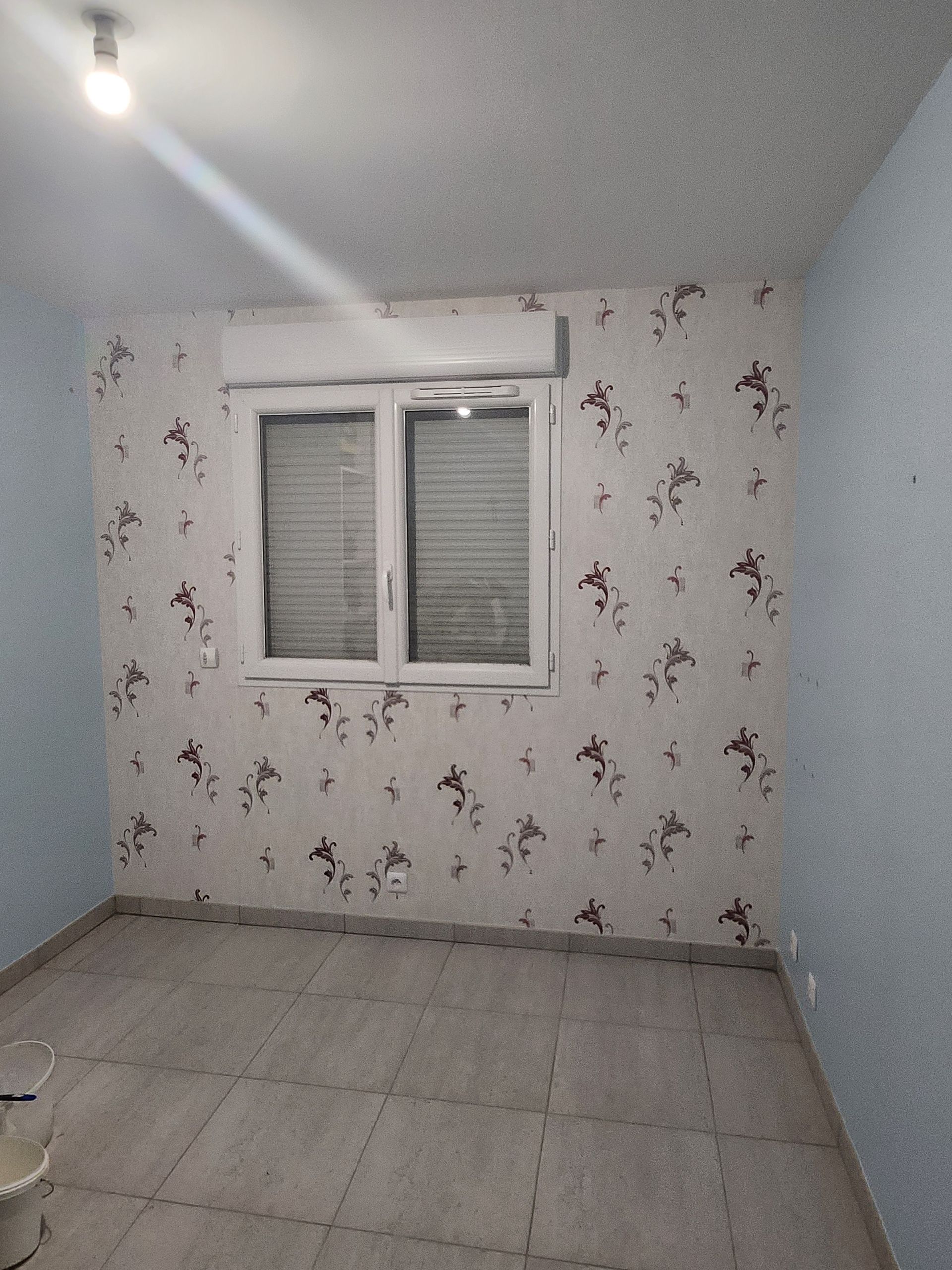 Une pièce vide aux murs bleu clair, un mur d'accent à motifs floraux, une fenêtre centrale avec stores et un sol carrelé.