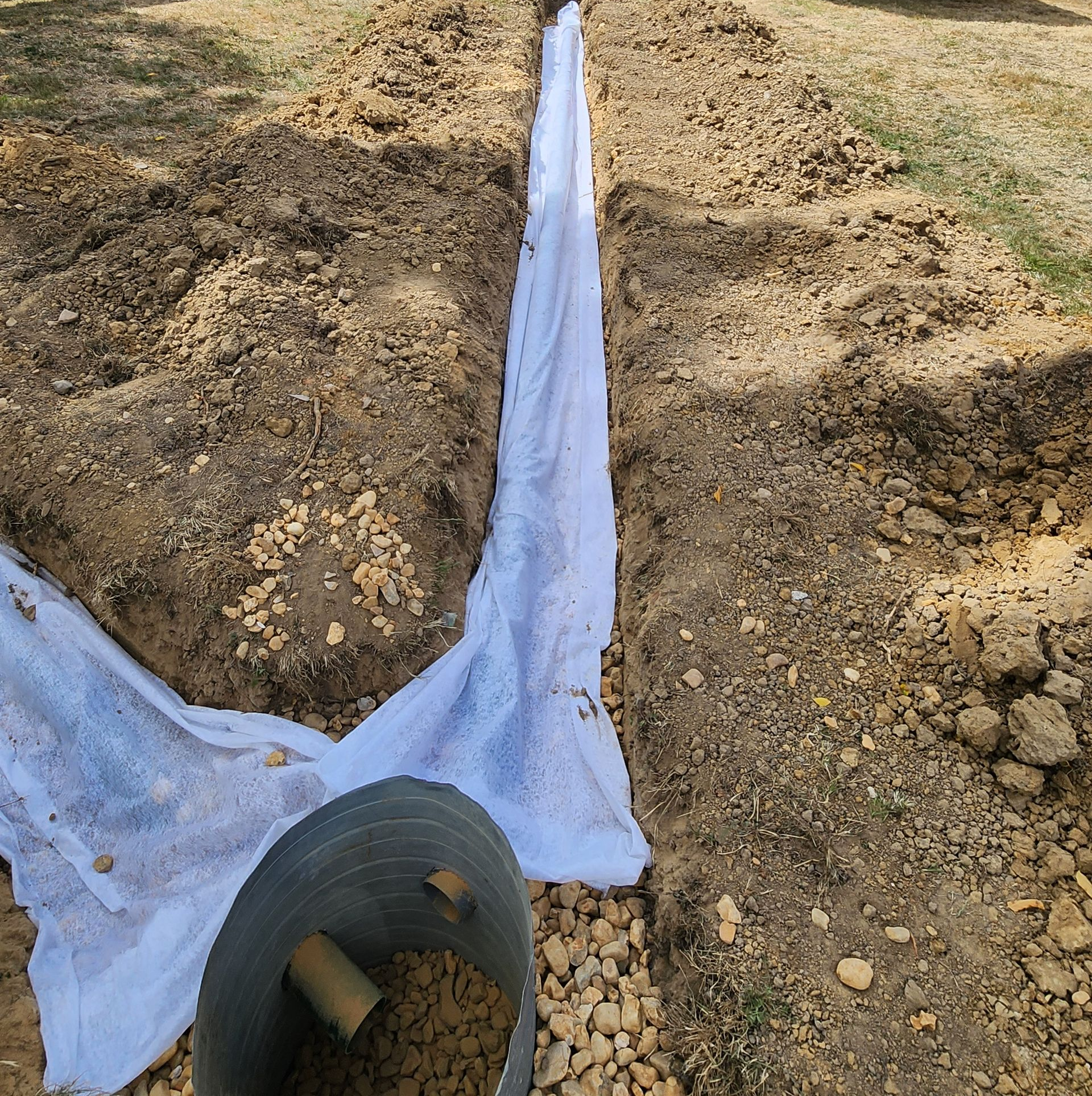 Une tranchée tapissée de géotextile blanc pour un drain français, avec un aménagement extérieur et un camion utilitaire à l'arrière.